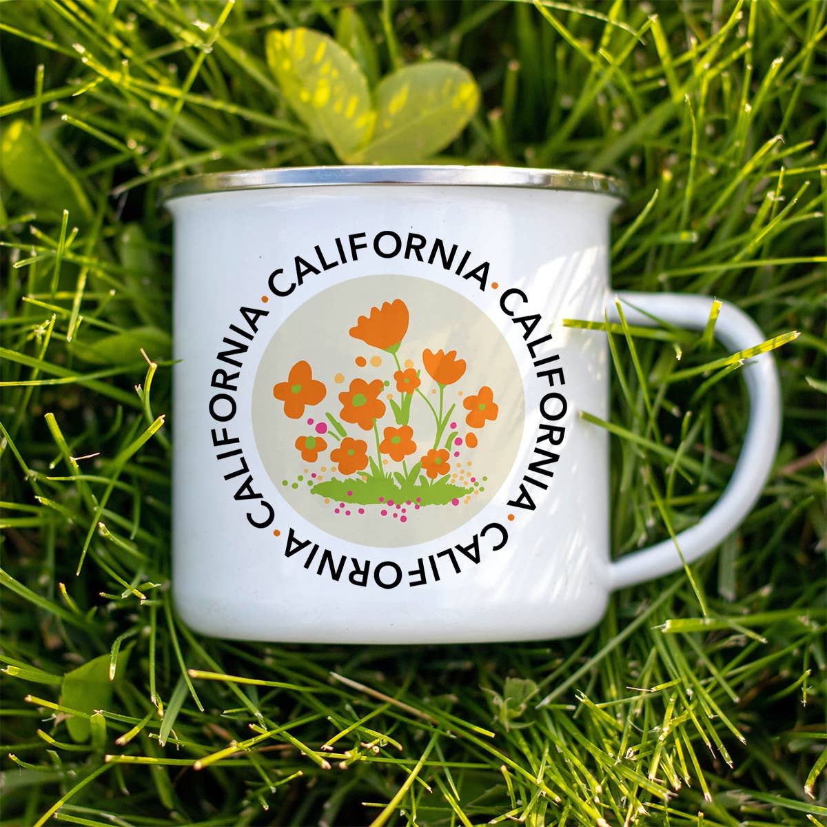 A white enamel camp mug with a silver rim, featuring a California poppy design and the word 'CALIFORNIA' repeated around the center, photographed outdoors on green grass.