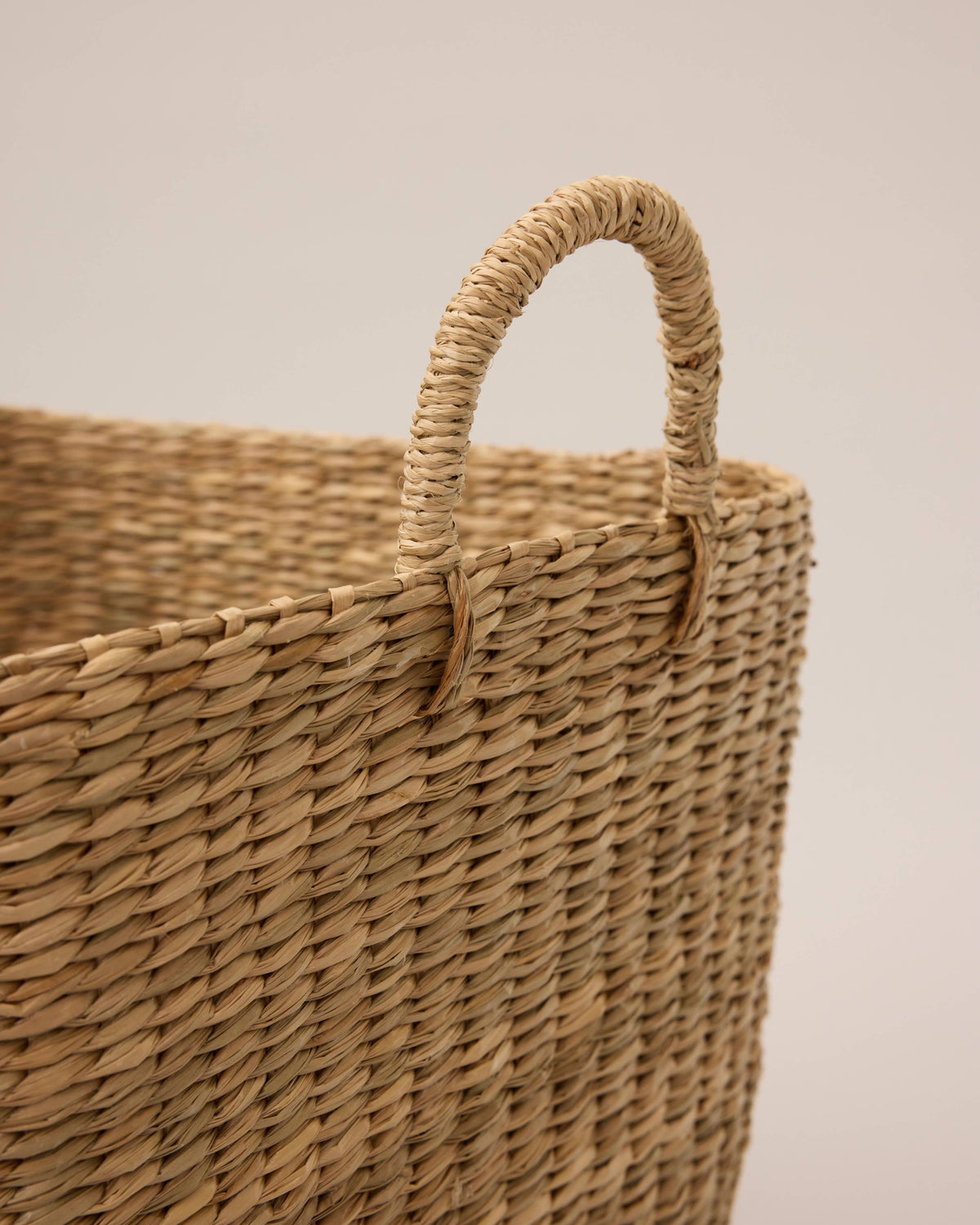 Close-up of a woven natural fiber basket with a single loop handle, showing the textured straw material in a warm tan color against a plain light background.