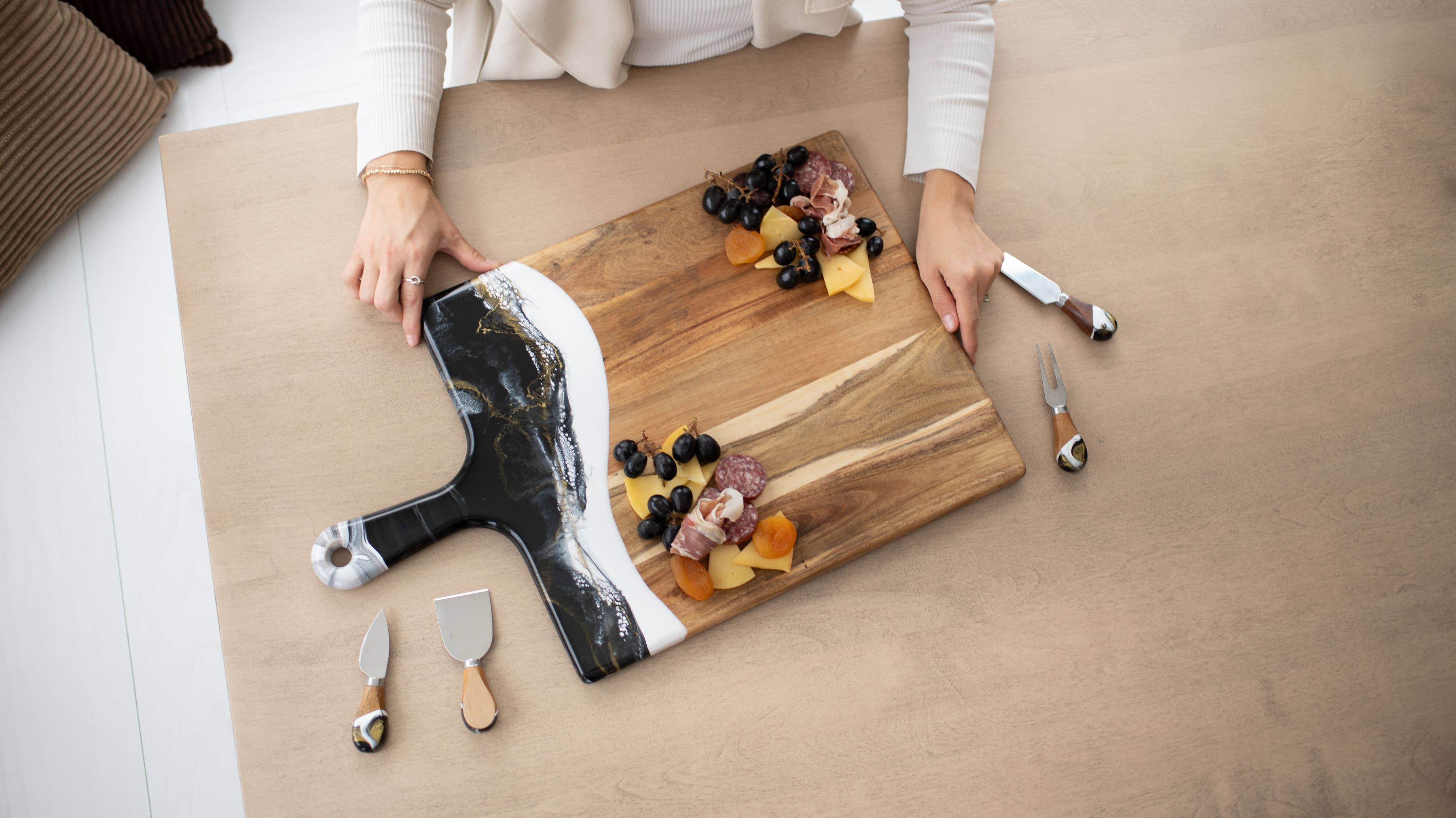 Person arranging food on a wooden cutting board with kitchen tools on a light wood floor.