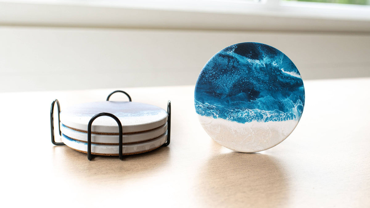 A set of round coasters on a light wood tabletop near a window. One coaster stands upright on the right, showing a blue ocean wave pattern over a white base.