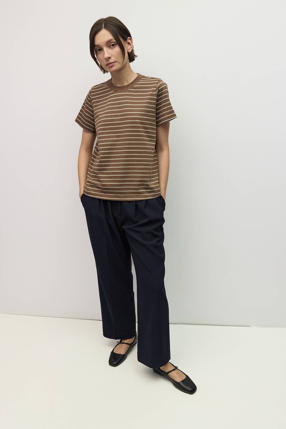 A model stands against a plain light gray studio wall wearing a brown short-sleeve crewneck t-shirt with thin horizontal beige stripes, dark navy wide-leg trousers, and black Mary Jane flats.