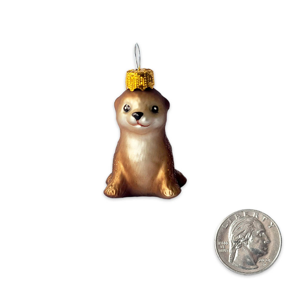 A small brown and cream otter Christmas ornament with a gold glittery cap, shown on a white background with a US quarter coin in the lower right for scale.