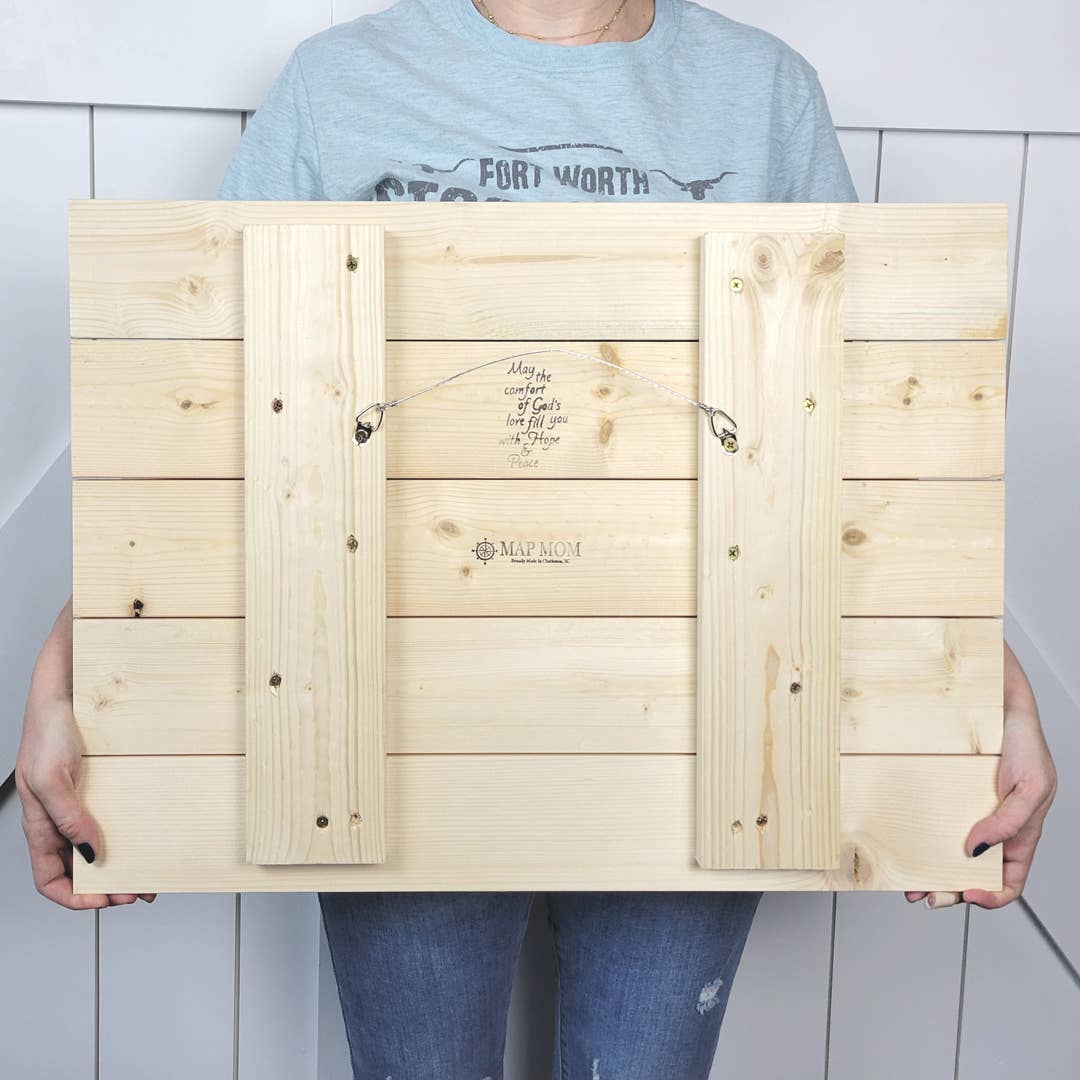 A person holding a light natural wood plank sign or panel with two vertical support boards and a hanging wire. The wood has small printed text and logo in the center.