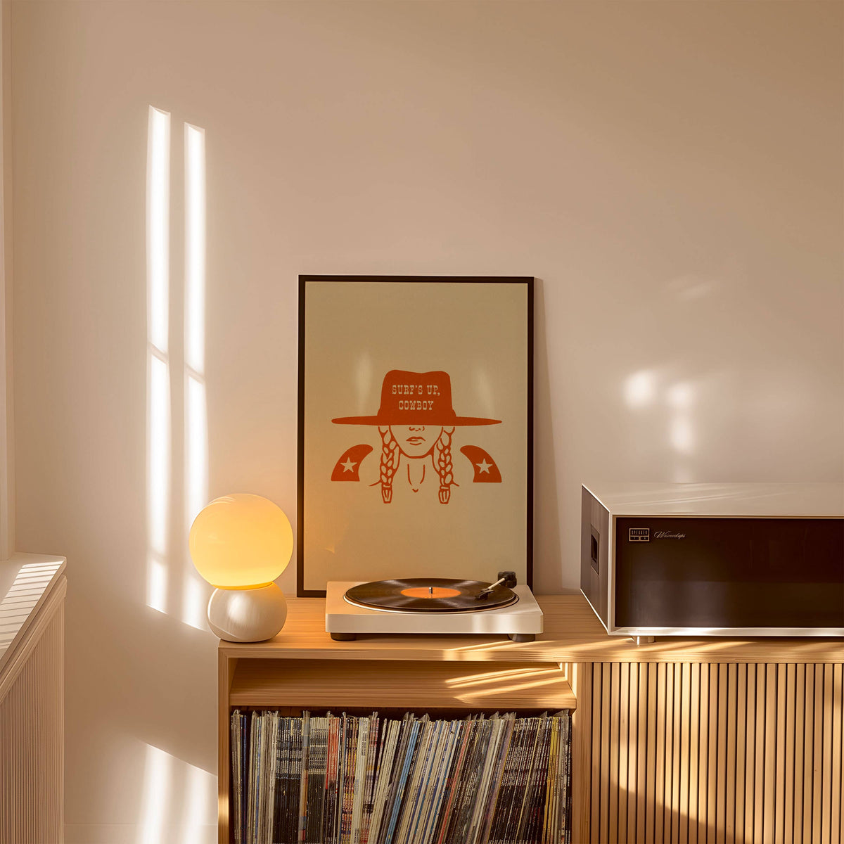 Record player on a wooden cabinet with vinyl records, framed art, and a lamp in a cozy room.