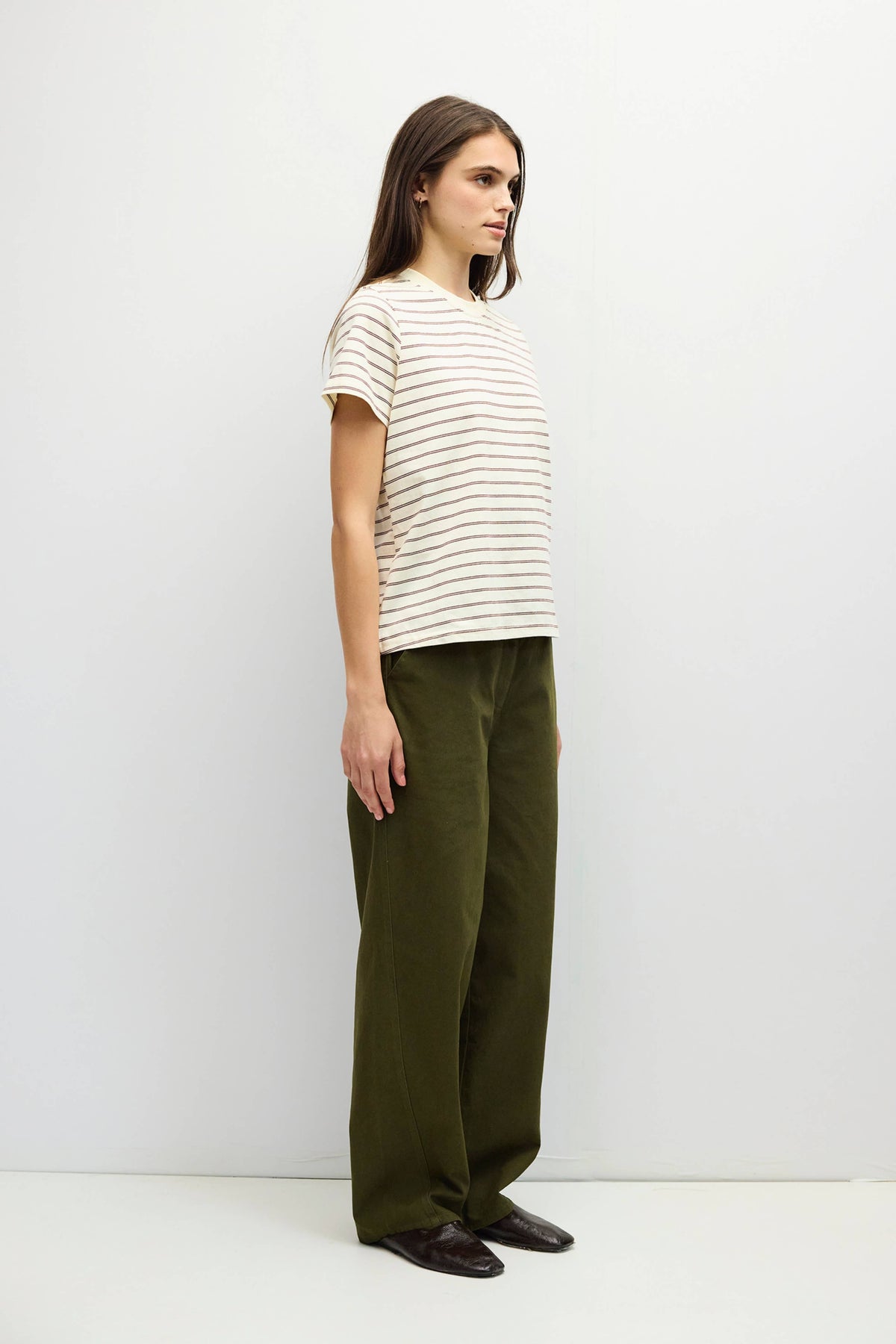 A woman stands in profile wearing a cream short-sleeve t-shirt with thin dark horizontal stripes and olive green wide-leg trousers, paired with dark brown shoes, against a plain light gray studio background.