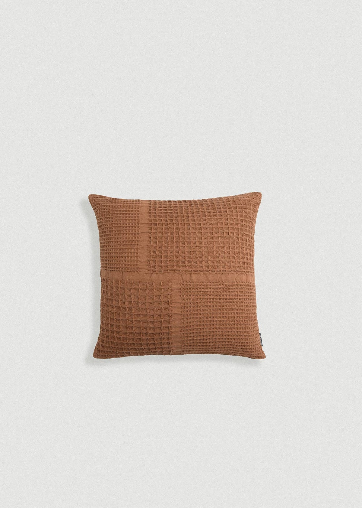 A square throw pillow in a warm terracotta brown color, featuring a textured waffle-weave pattern, shown against a plain light gray background.