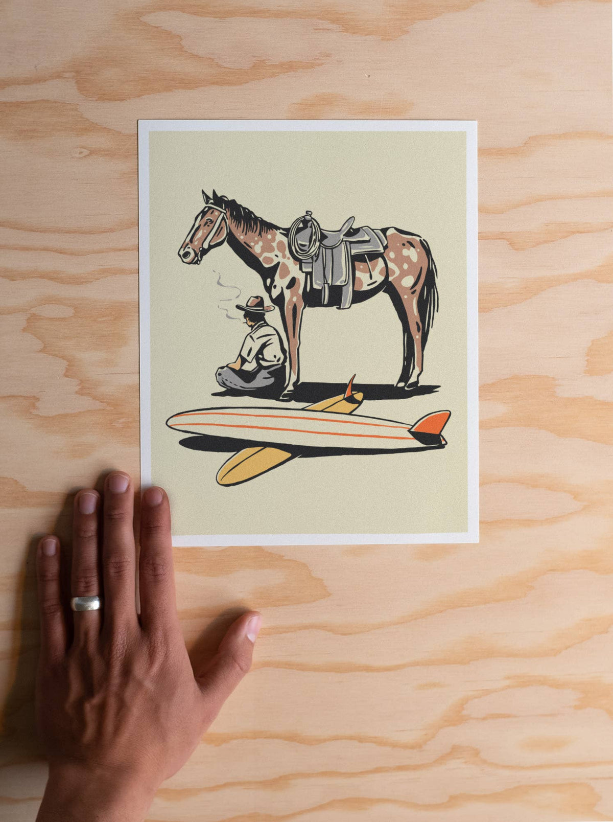 A square art print featuring an illustrated horse, rider, and surfboard, placed on a light wood surface with a hand holding the left edge for scale.