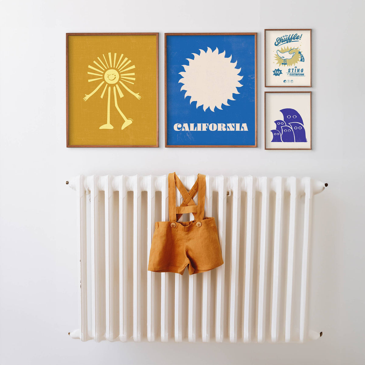A styled interior wall with four framed art prints above a white radiator. A small mustard-yellow garment hangs from the radiator.