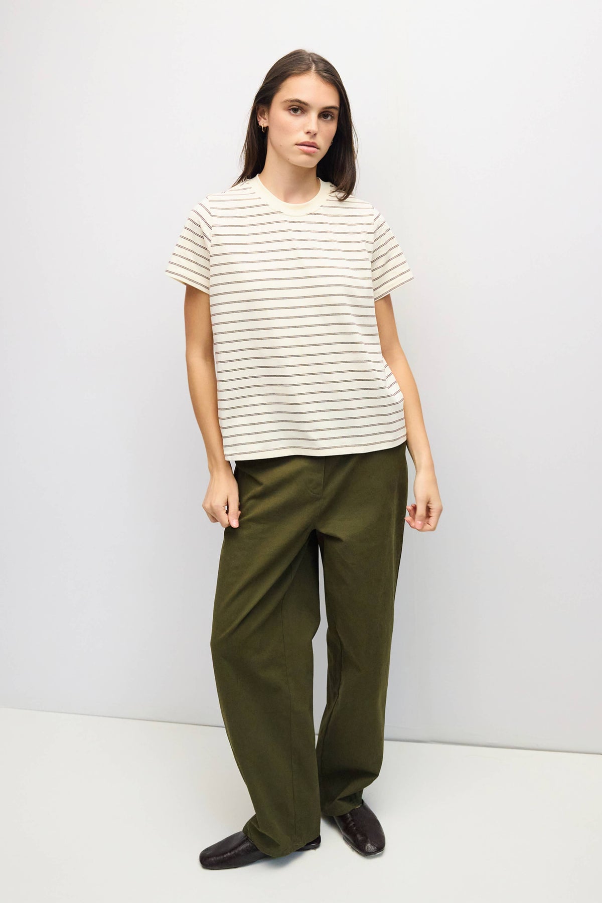 A woman modeling a short-sleeve crewneck t-shirt with thin dark horizontal stripes on an off-white base, paired with olive green wide-leg trousers and black shoes, against a plain light gray studio background.