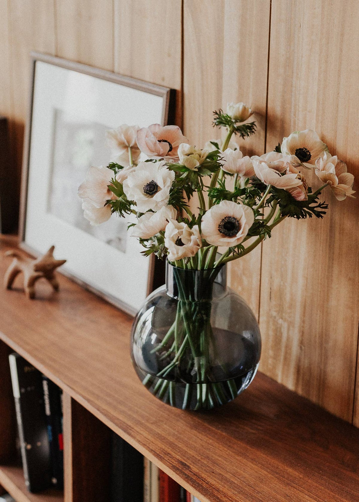 A bouquet of pale blush and cream anemone flowers with green stems arranged in a round dark-tinted glass vase, placed on a wooden shelf against a wood-paneled wall with a framed print nearby.
