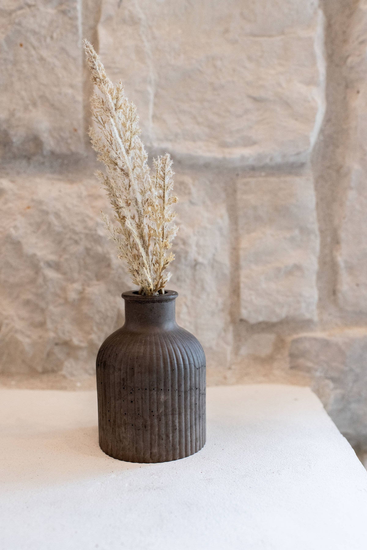 Black textured vase with dried plants on a white surface against a stone wall background