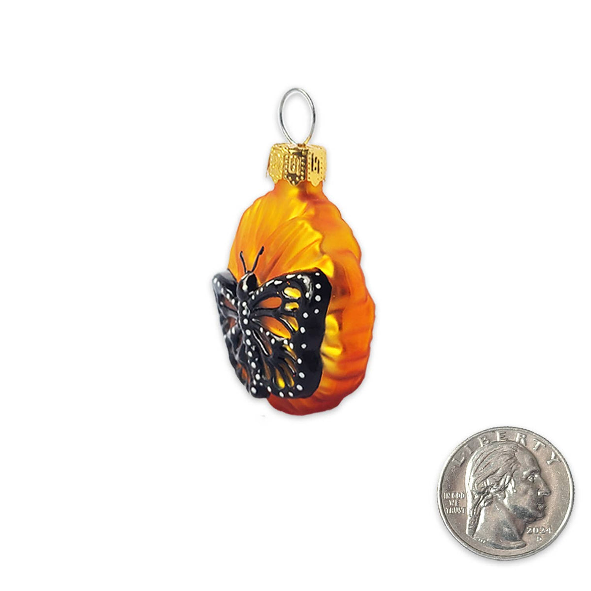 An orange and yellow glass ornament shaped like a pumpkin, featuring a black and orange butterfly decoration, shown next to a US quarter coin for scale on a white background.