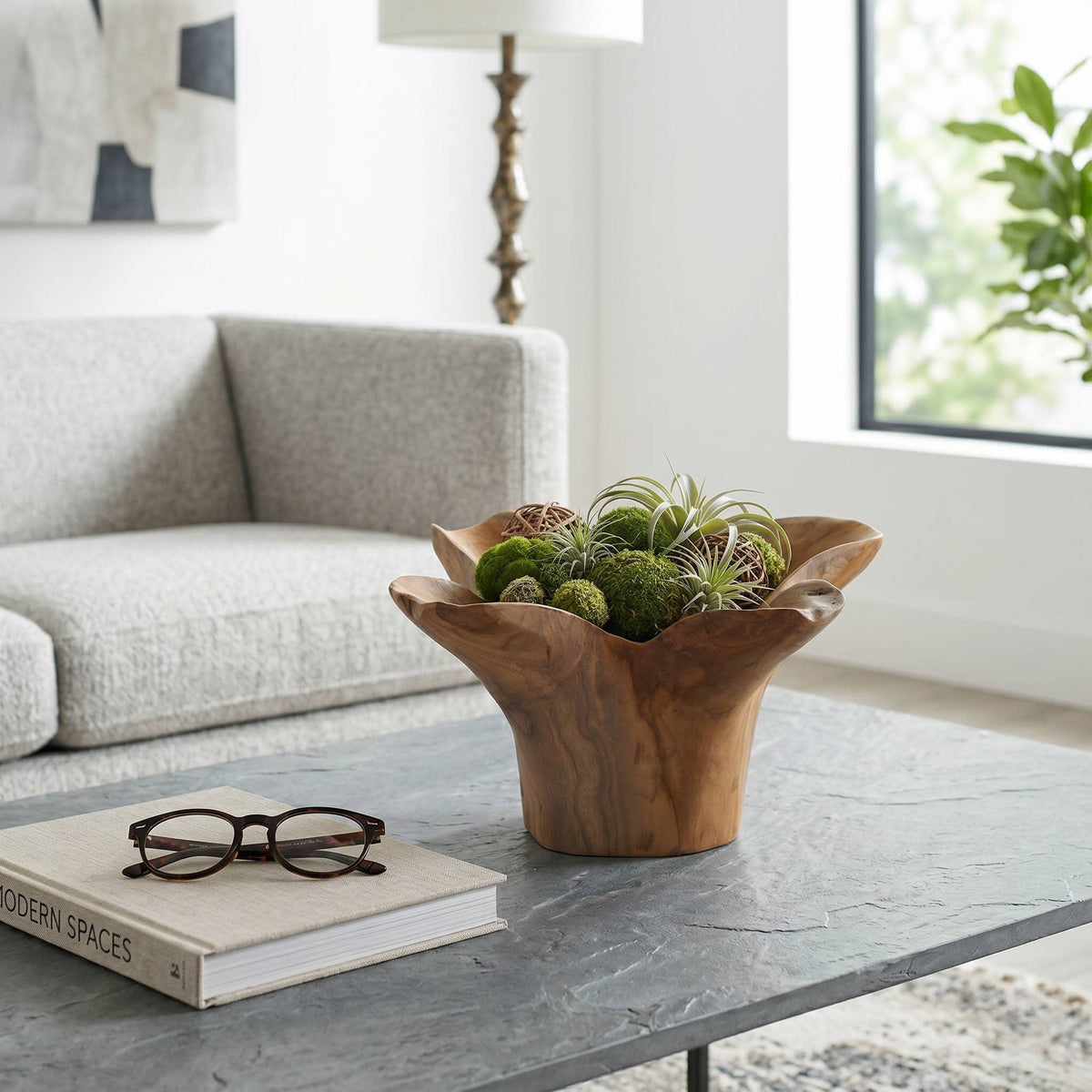 A rustic wooden bowl planter filled with green succulents and air plants, styled on a gray marble coffee table in a modern living room setting.