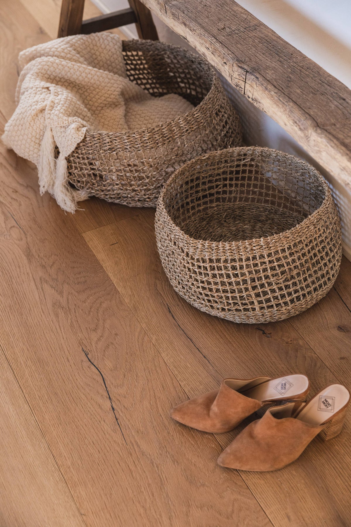Two round woven jute baskets on a light wood floor, with a cream knit throw draped over one basket and a pair of tan pointed-toe mule sandals nearby.