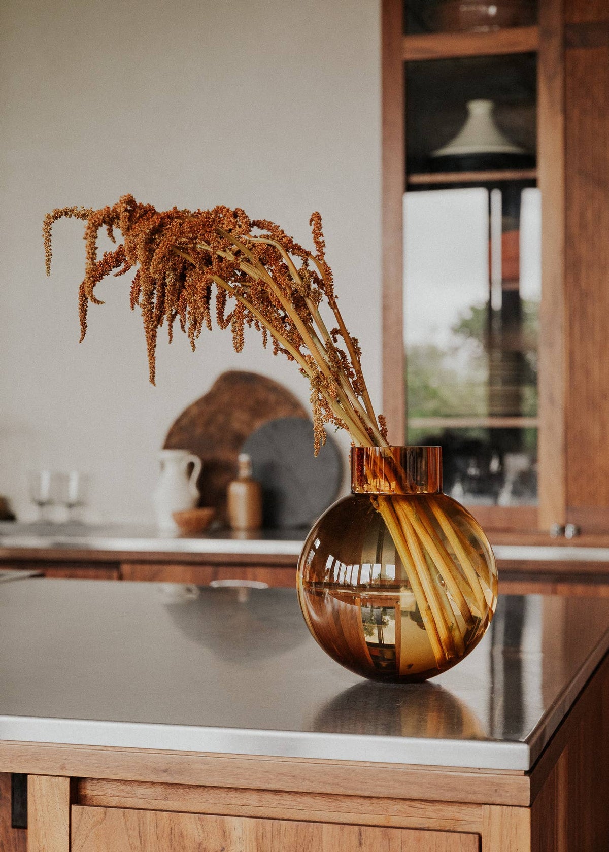 An amber-brown glass vase with a matching cylindrical top, filled with dried ornamental grass stems, set on a dark countertop in a warm wood kitchen interior.