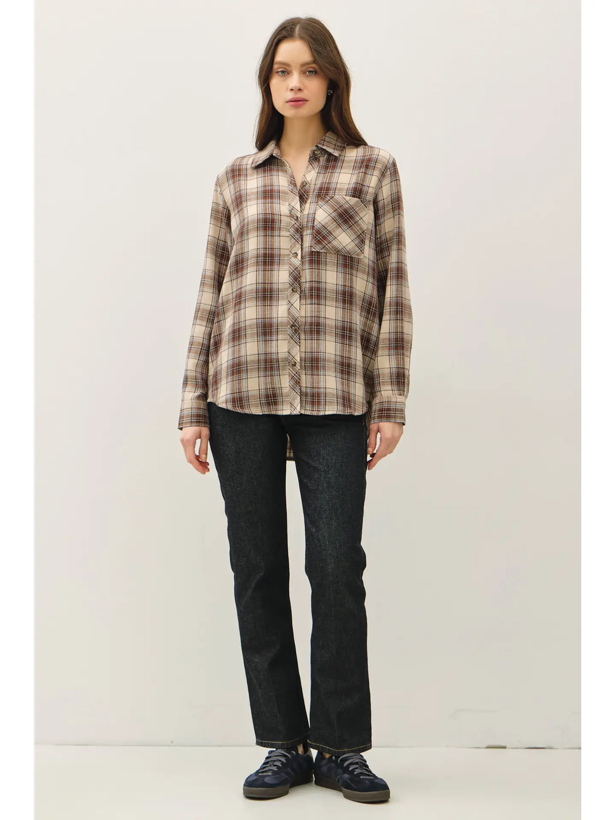 A woman stands against a plain light background wearing a long-sleeve plaid button-up shirt in beige, brown, and black tones, paired with dark jeans and dark shoes.
