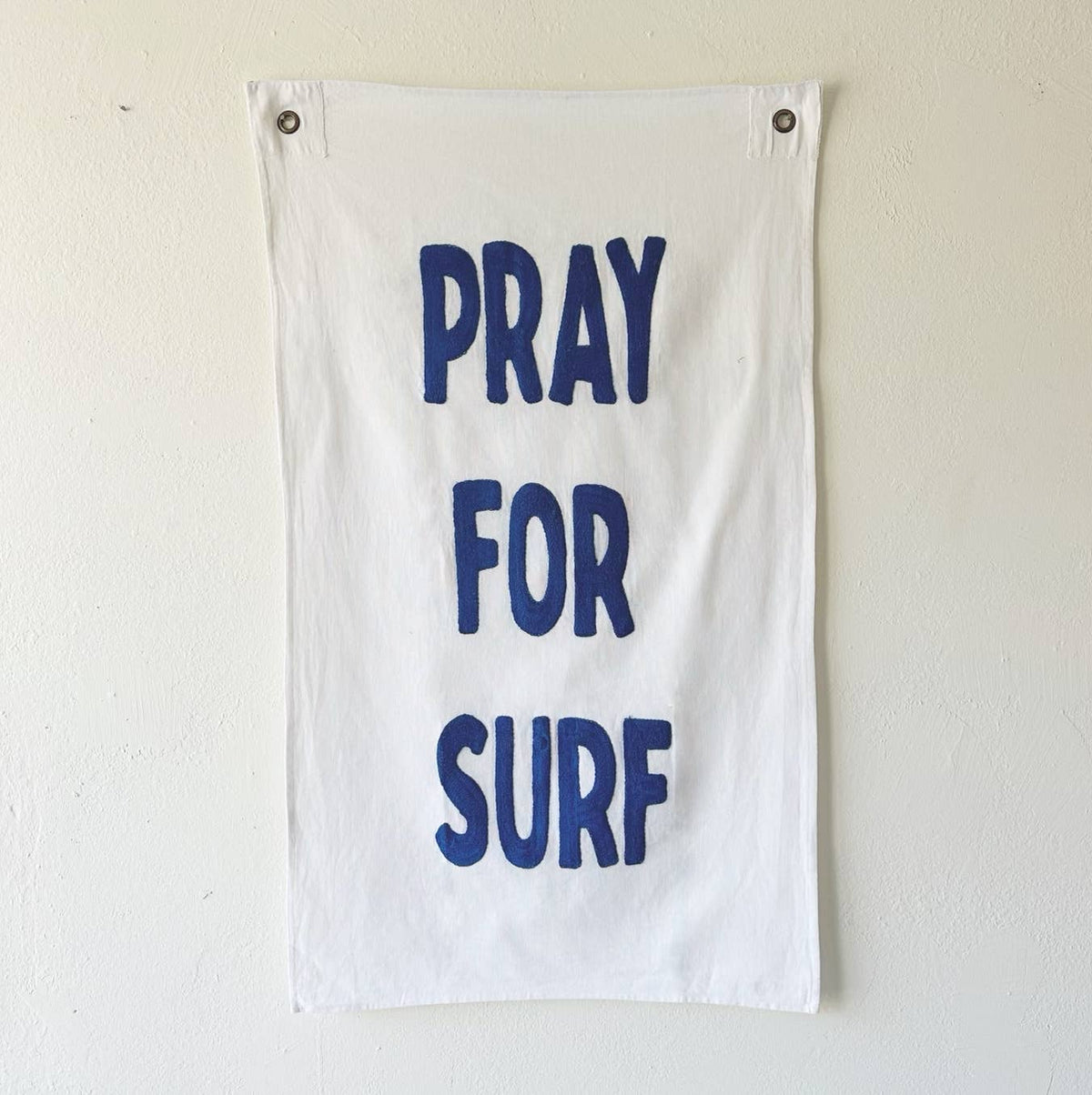 A white fabric wall hanging or towel with the phrase 'PRAY FOR SURF' printed in large blue letters, hanging on a light beige wall with two metal grommets at the top corners.