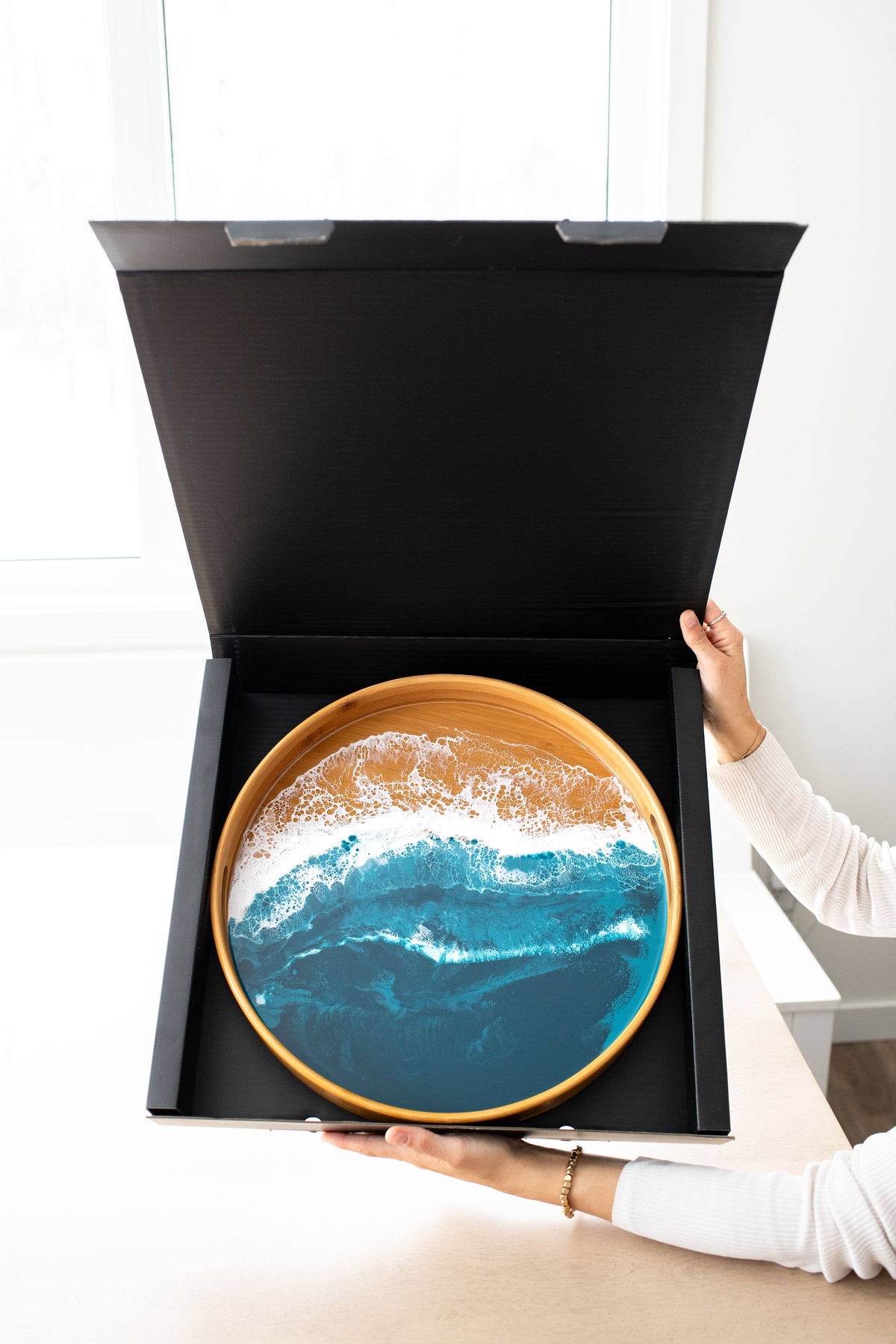 A round decorative tray with a gold rim and an ocean wave design in shades of blue, white, and tan, displayed inside an open black gift box being held by a person.