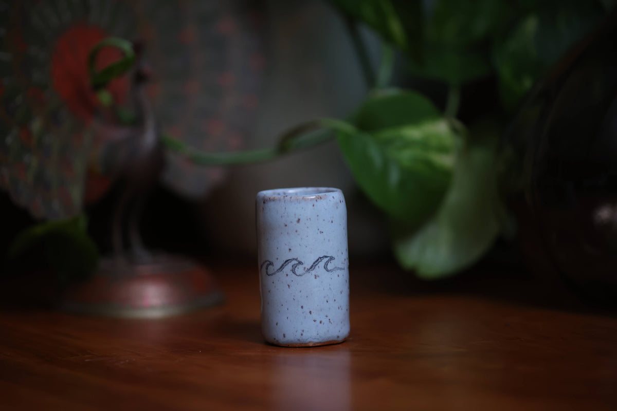 Small cylindrical ceramic object with wavy line design on a wooden surface with plants in the background