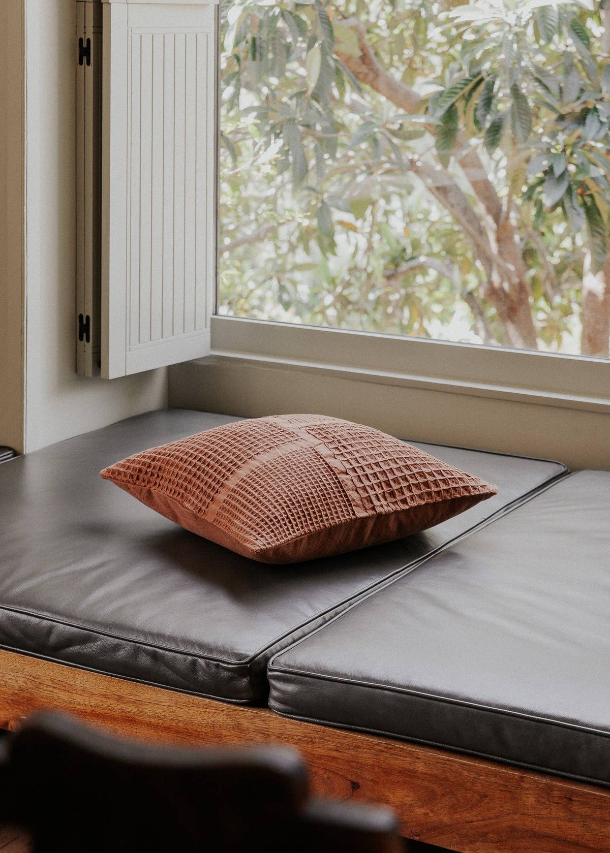 A square waffle-weave throw pillow in a dusty terracotta pink color rests on dark gray leather bench cushions positioned by a window with a view of green trees outside.