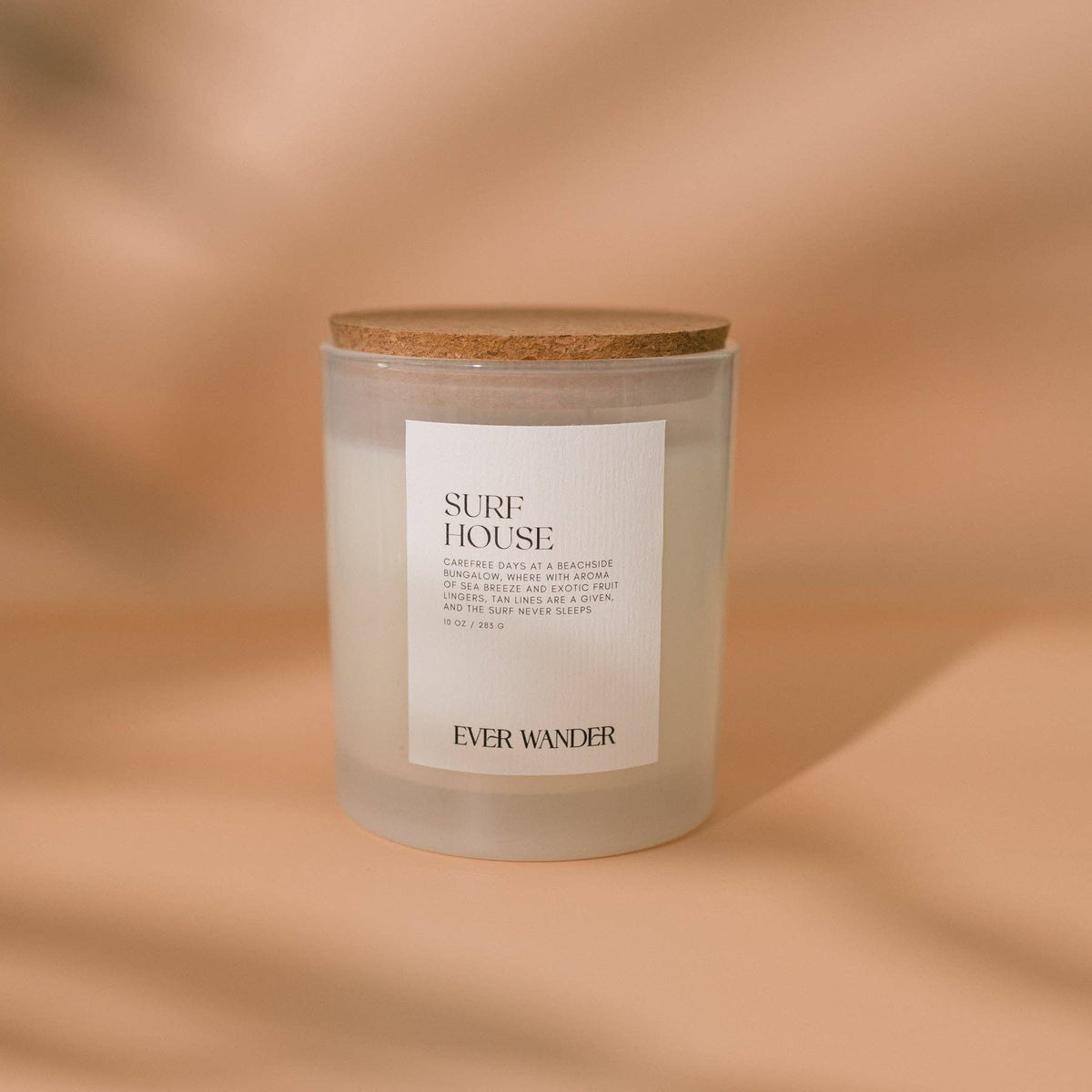 A frosted glass candle jar with a natural cork lid and a white label reading 'SURF HOUSE' and 'EVER WANDER', set against a warm beige background with soft shadows.
