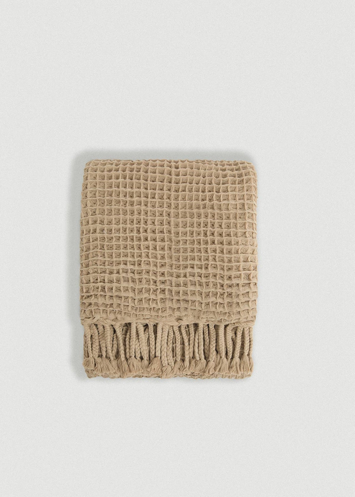 A folded beige woven throw blanket with a waffle texture and fringed edge, photographed on a light gray background.