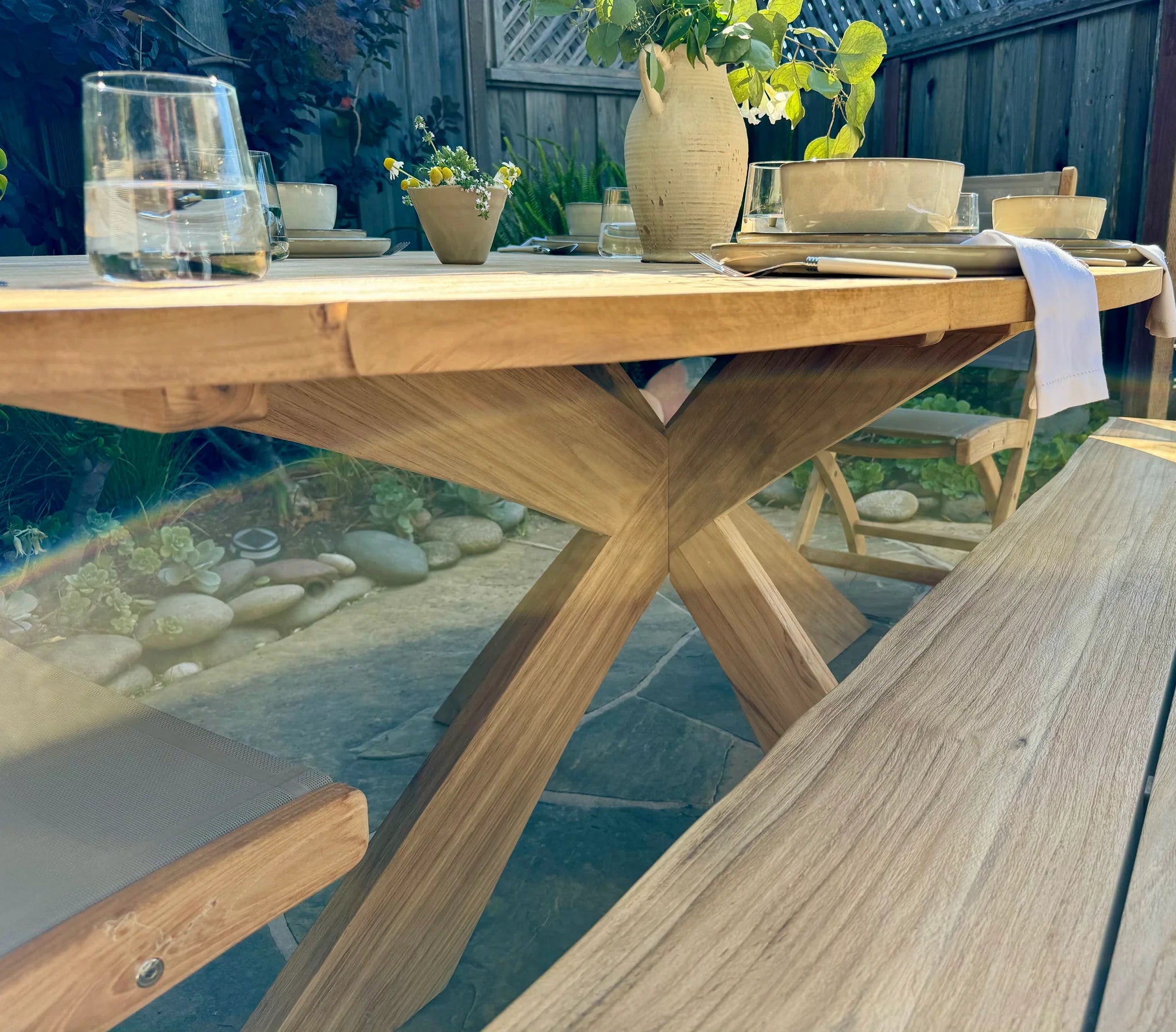 Wooden outdoor dining table with chairs and a vase of flowers on a patio.
