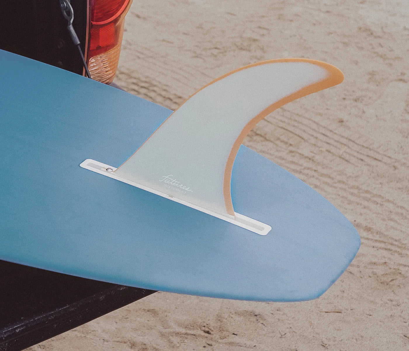 Close-up of a blue surfboard with a white and orange Futures Zack Flores Flex Fiberglass Single Fin (9.5") on a sandy background