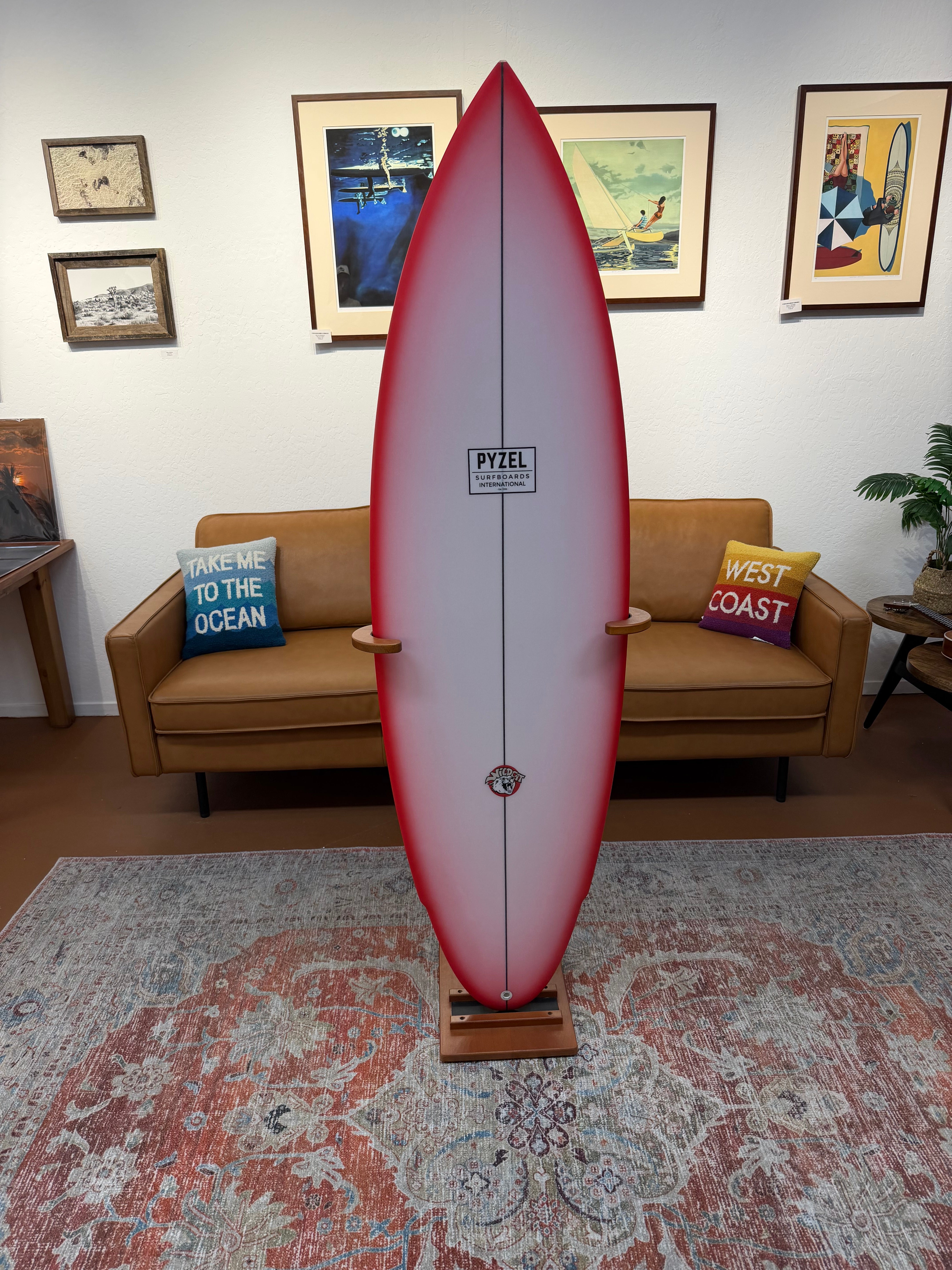 A vintage PTL surfboard with a red-to-white gradient and a black center stripe, displayed upright on a wooden stand in a living room setting.