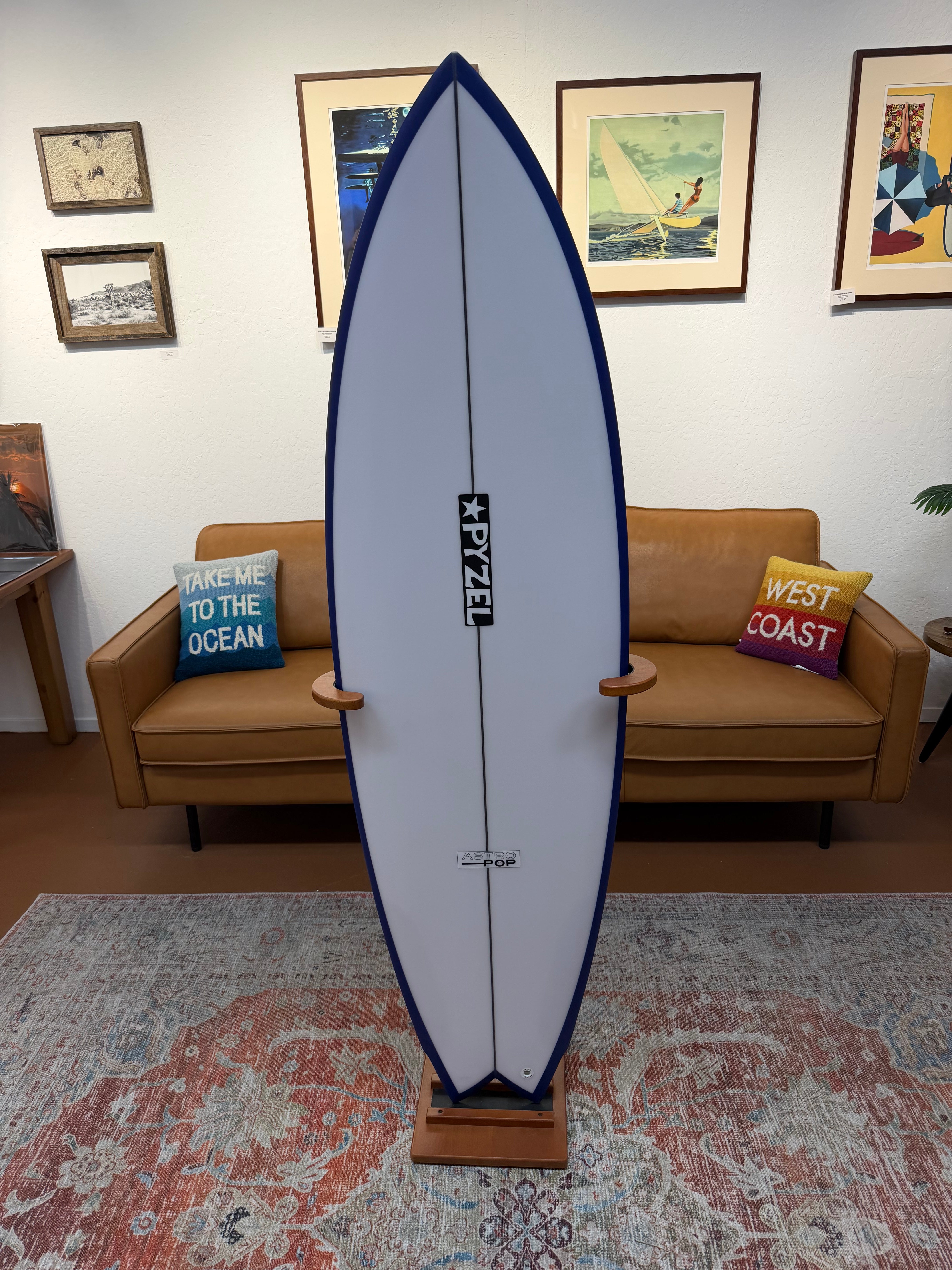 A Pyzel surfboard displayed vertically on a wooden stand in a living room setting, with a tan leather sofa, framed wall art, and a patterned rug in the background.