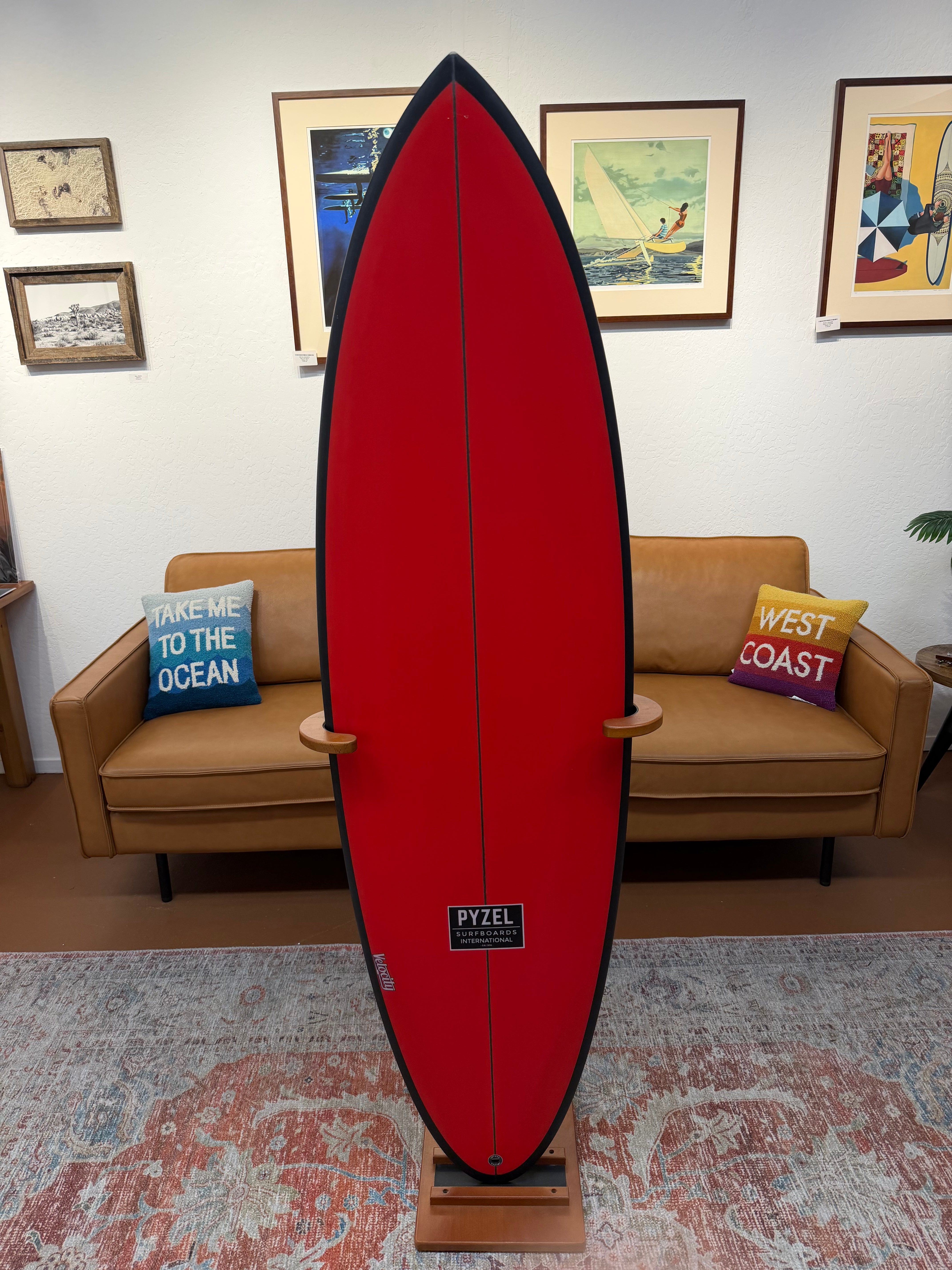 A red Pyzel surfboard displayed upright on a wooden stand in a living room setting, with a tan leather sofa, colorful throw pillows, and framed art on the wall behind it.