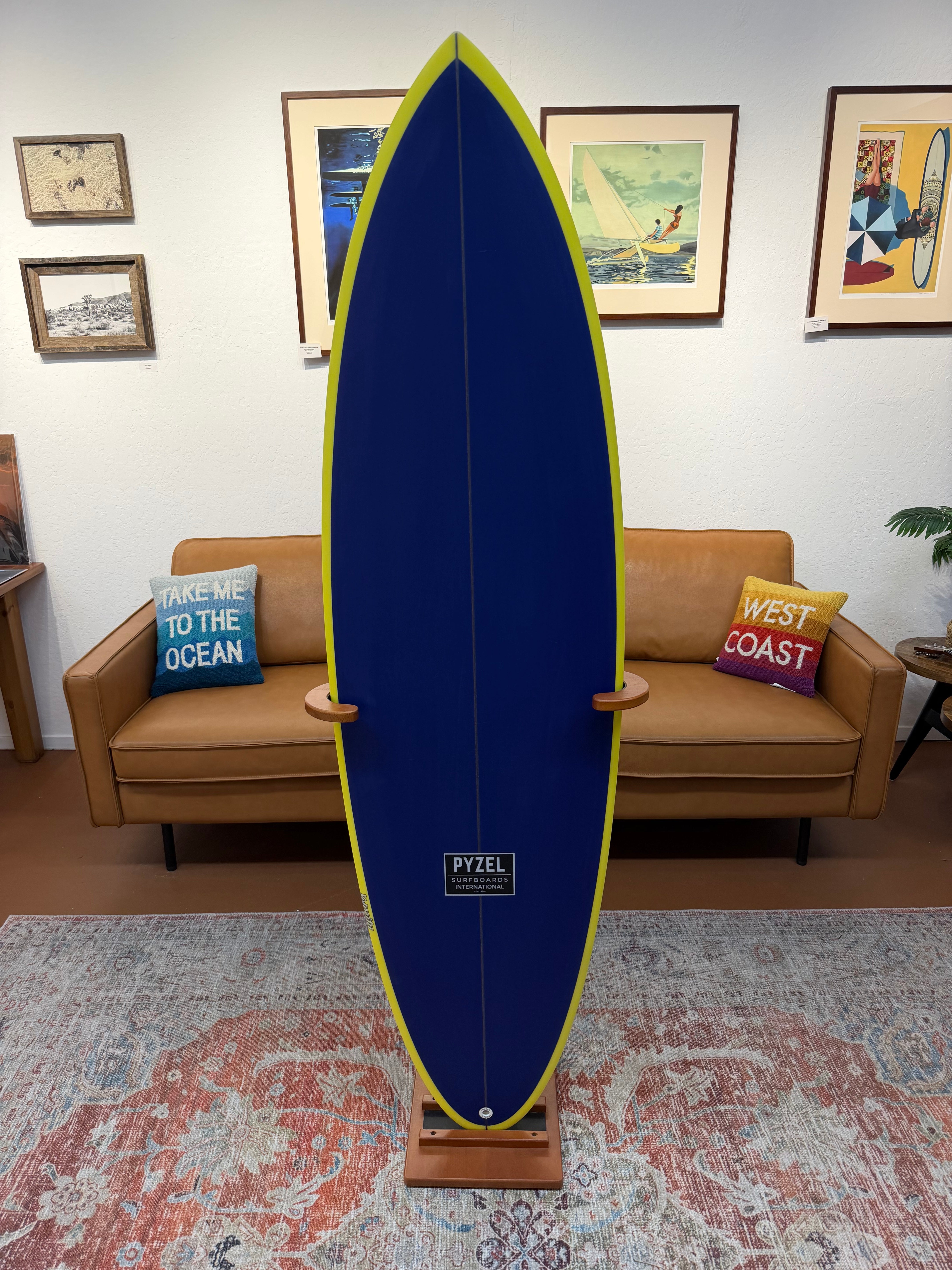 A long surfboard displayed upright on a wooden stand in a living room setting. The surfboard is dark blue with a yellow-green edge and a small black square logo near the tail.