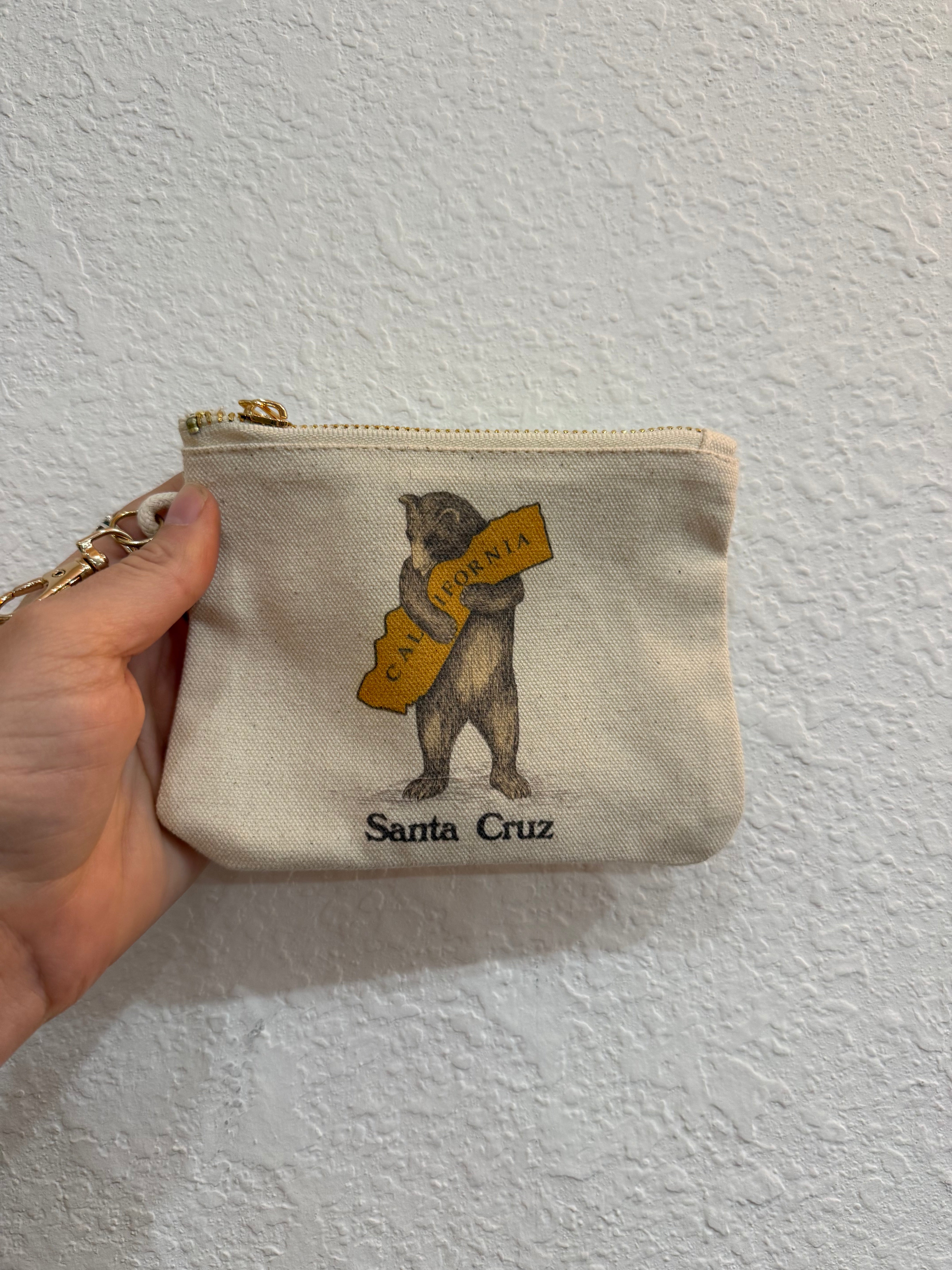 A hand holding a small canvas pouch with a gold zipper, featuring a vintage-style illustration of a bear holding a yellow sign that reads 'Santa Cruz' against a textured white wall.