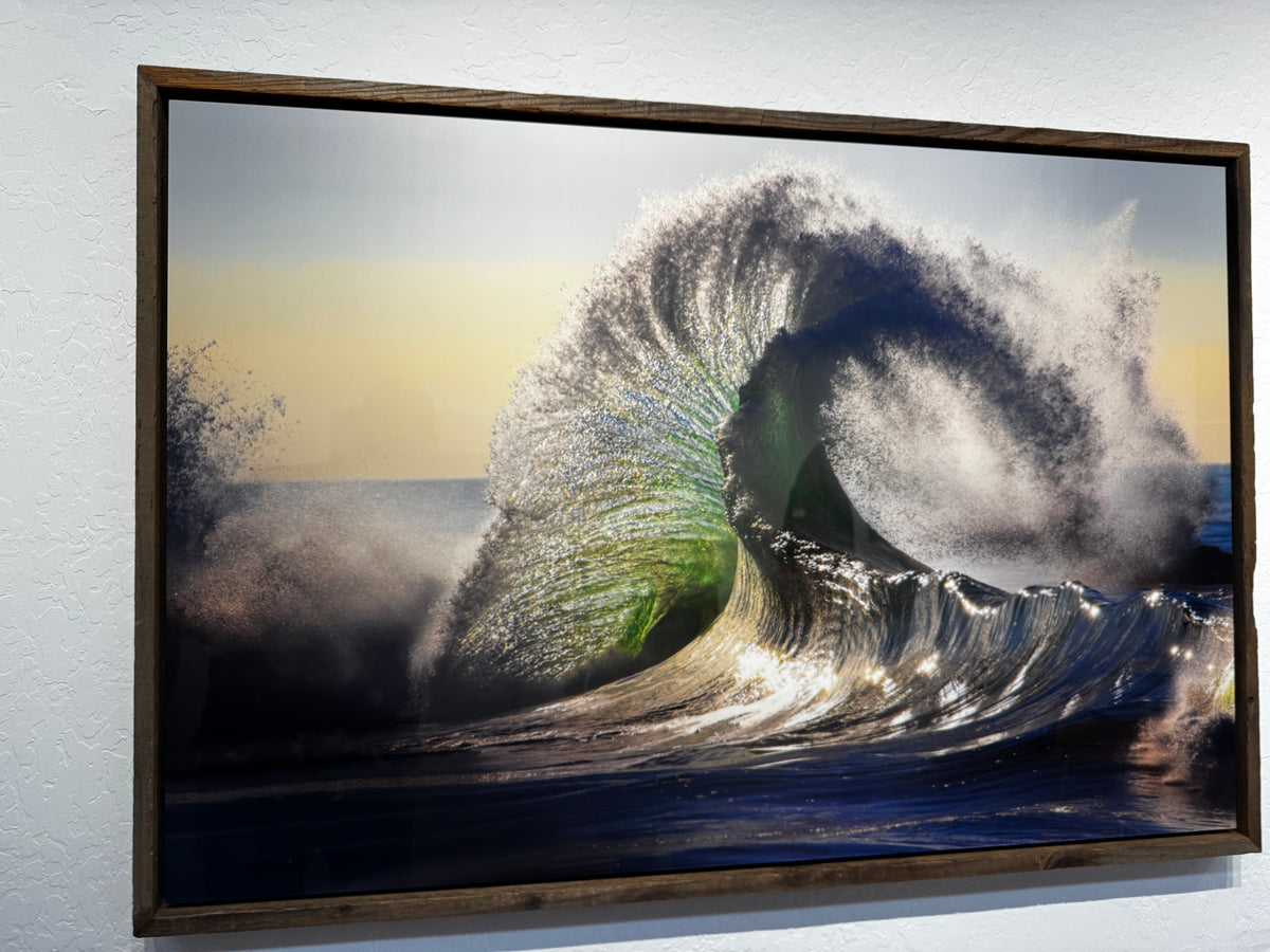 A framed photographic art print of a dramatic ocean wave curling, mounted on a textured white wall. The print features a dark brown wooden frame and depicts a wave with a vibrant green interior, white foam, and blue water under a pale sky.