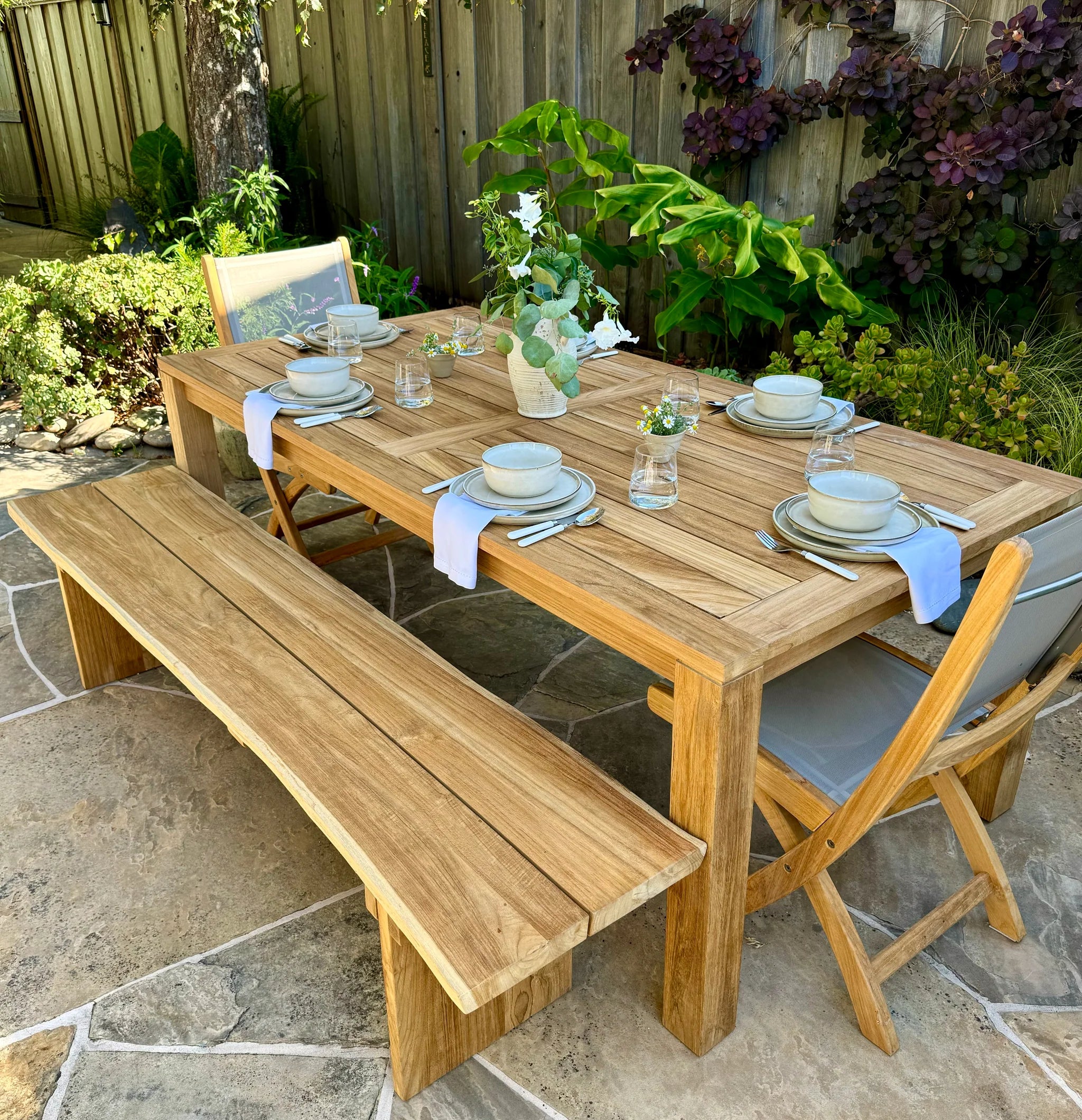 Wooden outdoor dining table set with chairs and a bench on a stone patio.