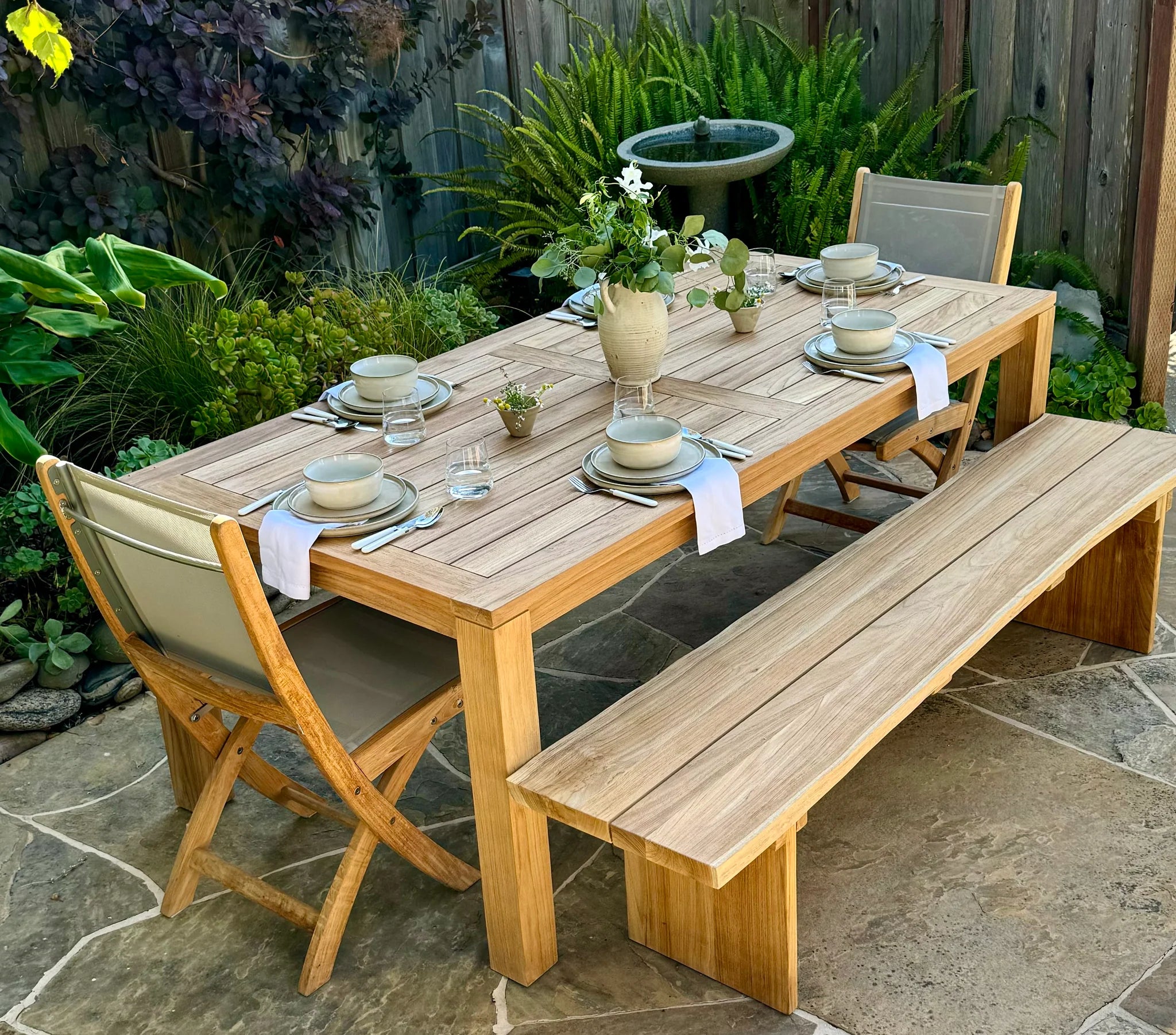 Wooden outdoor dining set with table, chairs, and bench on a stone patio surrounded by greenery.