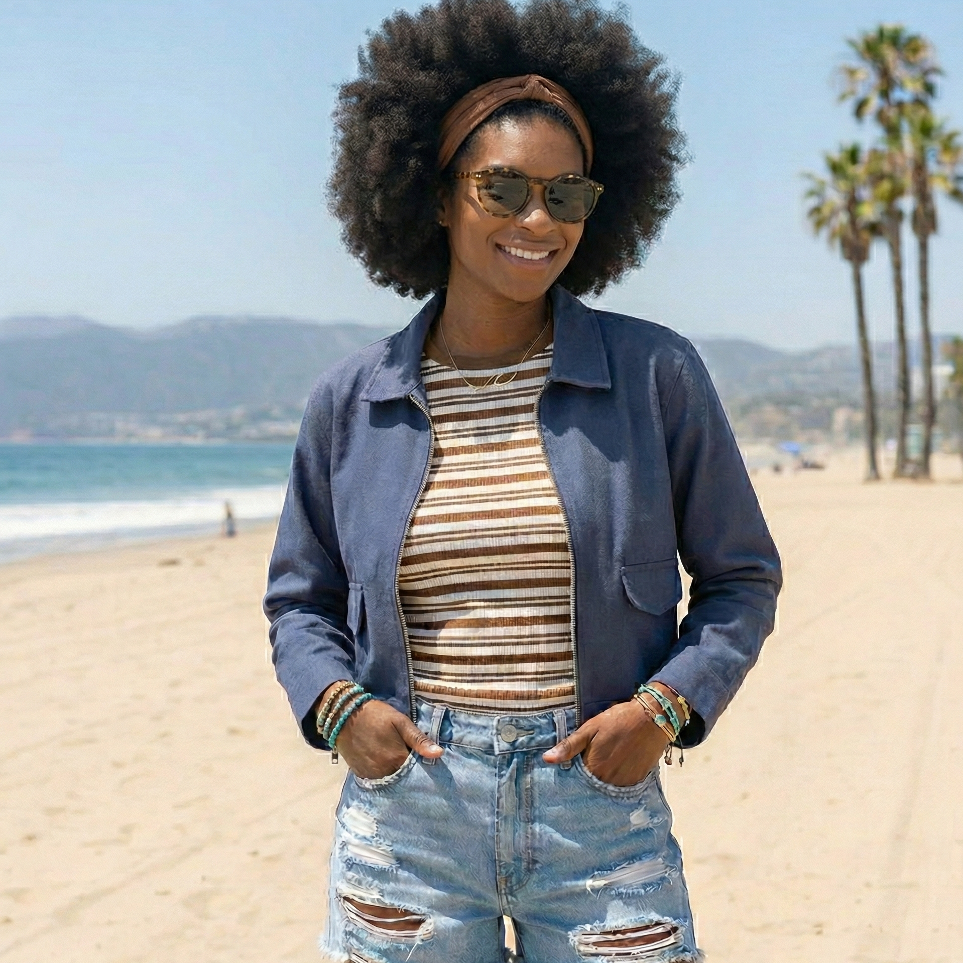 Woman on a beach wearing sunglasses, a blue jacket, striped shirt, and denim shorts.