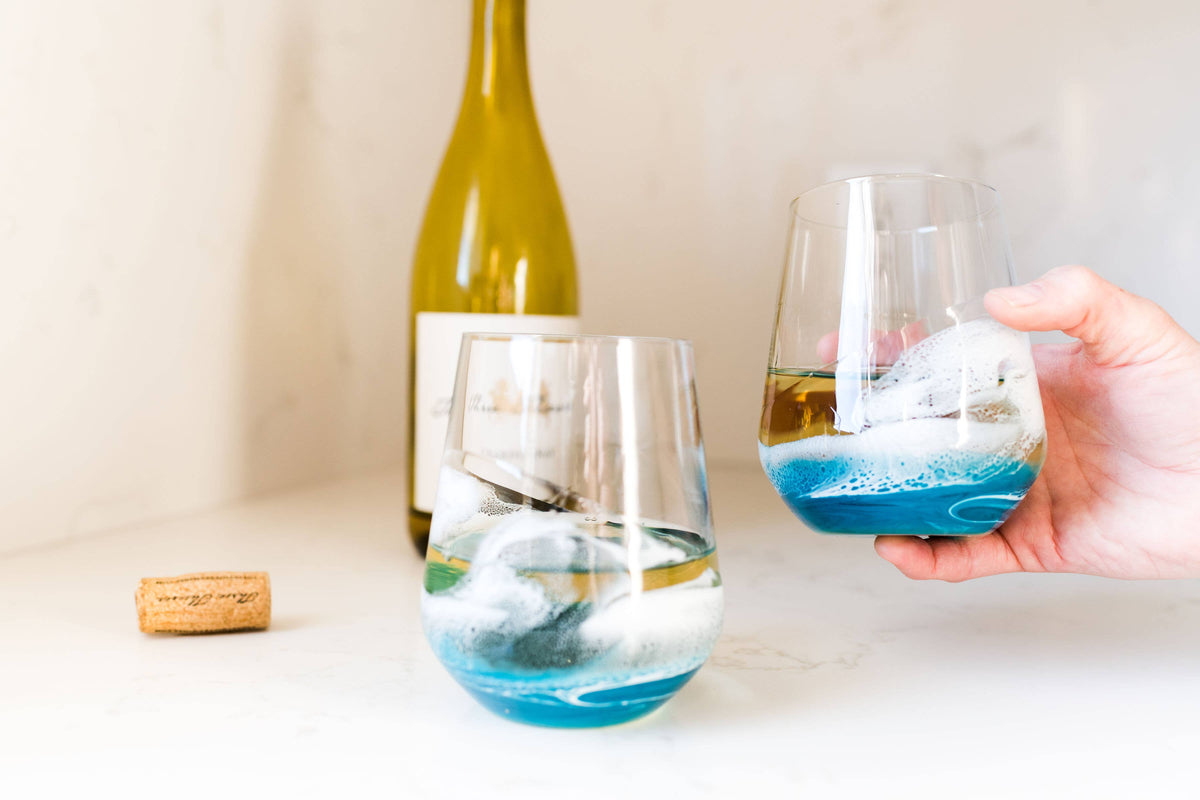Two wine glasses with blue and white designs, one being held by a hand, on a light surface with a bottle in the background.