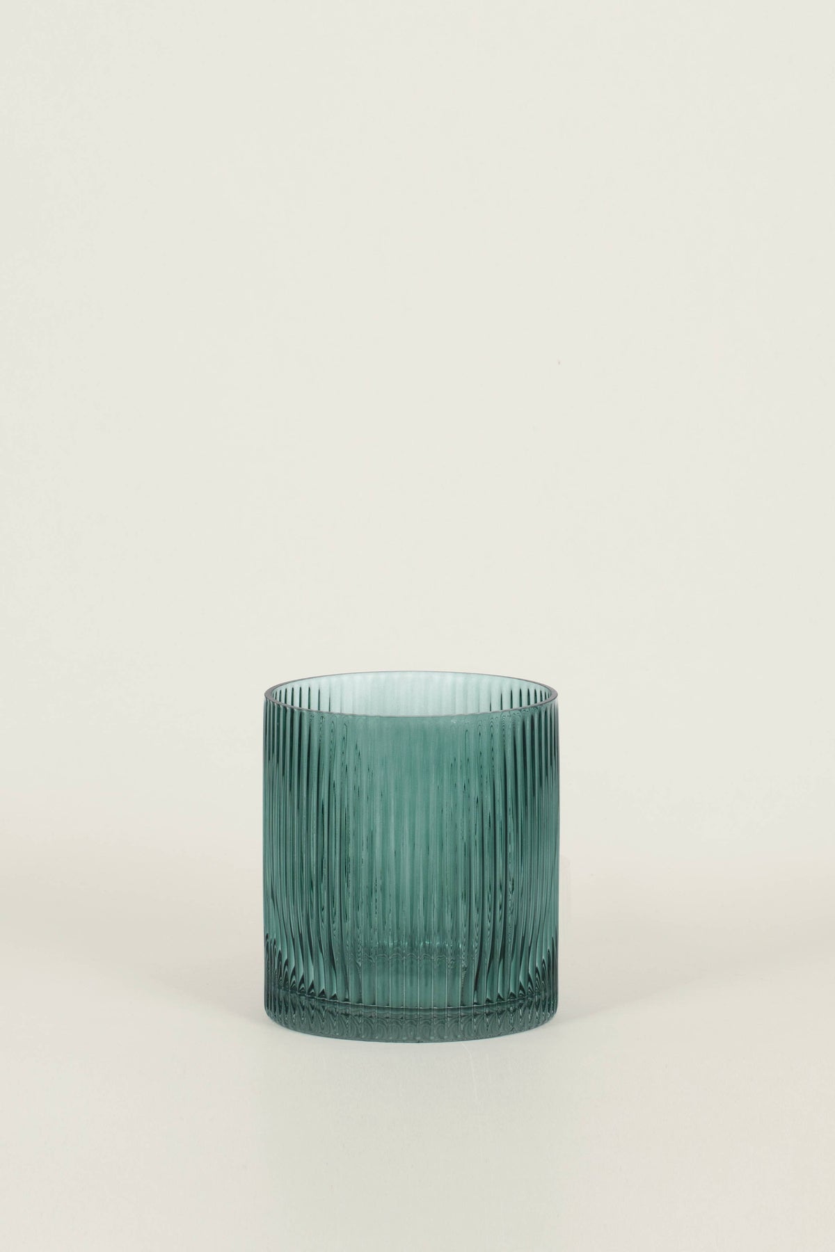 A small ribbed glass tumbler in a muted teal-green color, standing on a light beige surface against a matching light beige background.