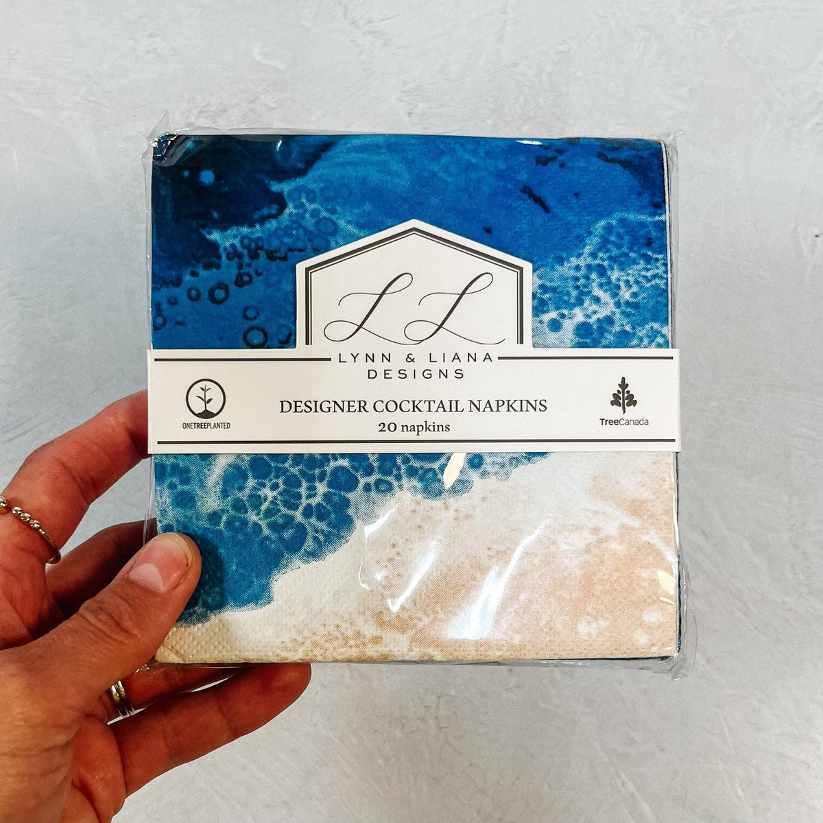 Pack of designer cocktail napkins with blue and beige design held by a hand on a light gray background.