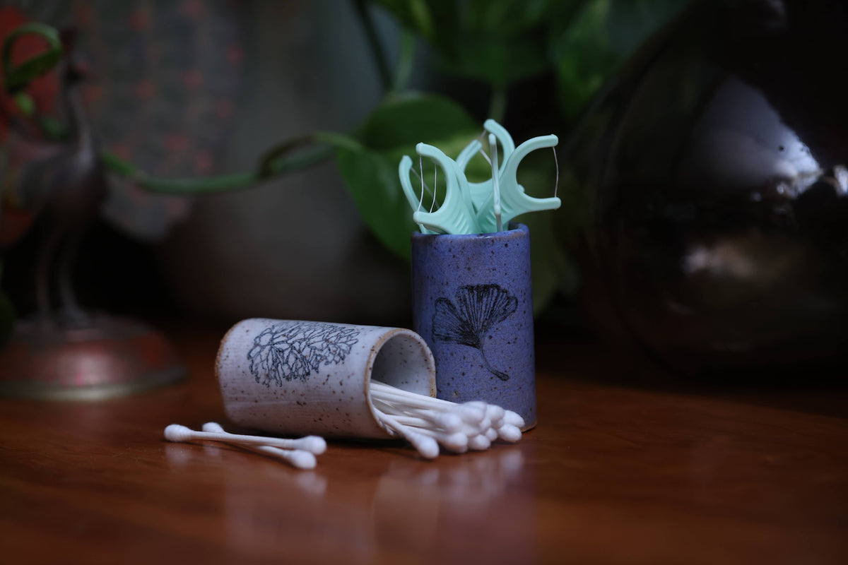 Two cylindrical containers with cotton swabs on a wooden surface, with a blurred natural background.
