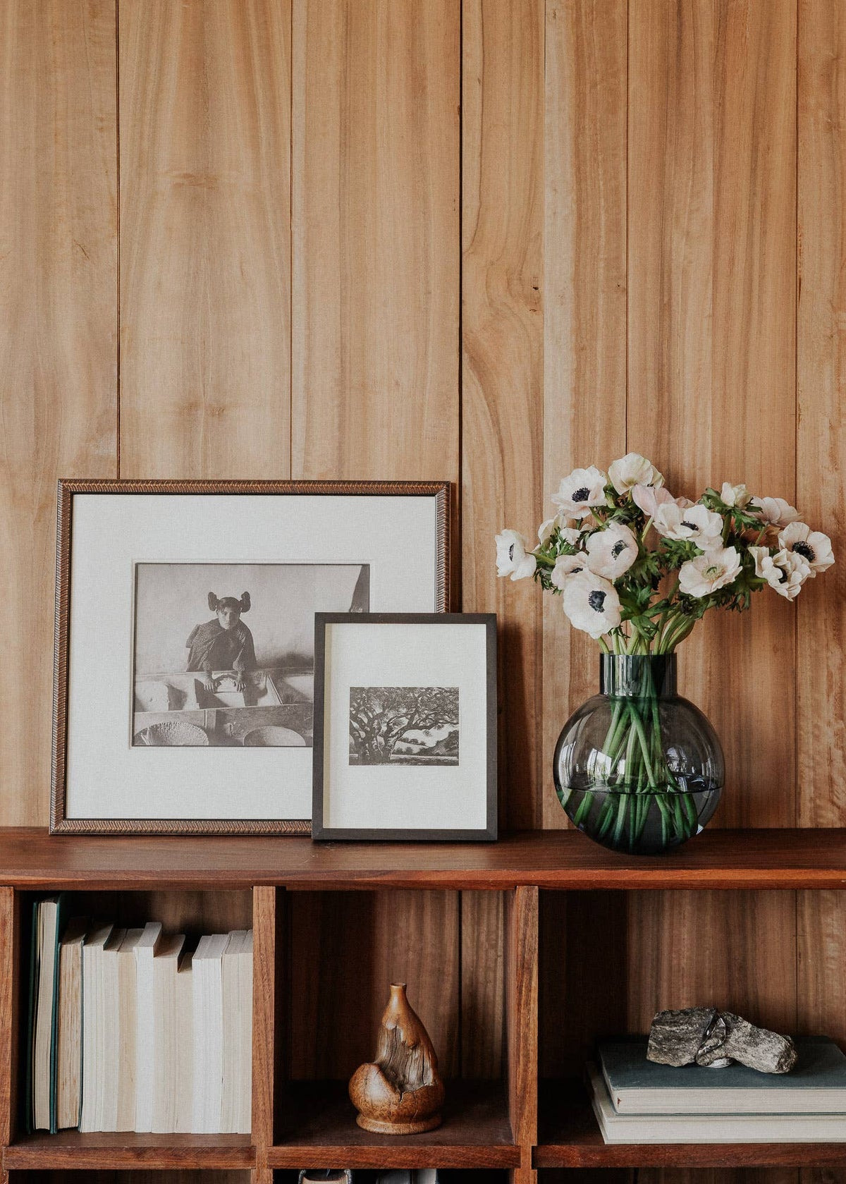 A styled interior scene with two framed black-and-white artworks leaning on a wooden shelf beside a green glass vase holding white flowers, with books and decor below against a wood-paneled wall.