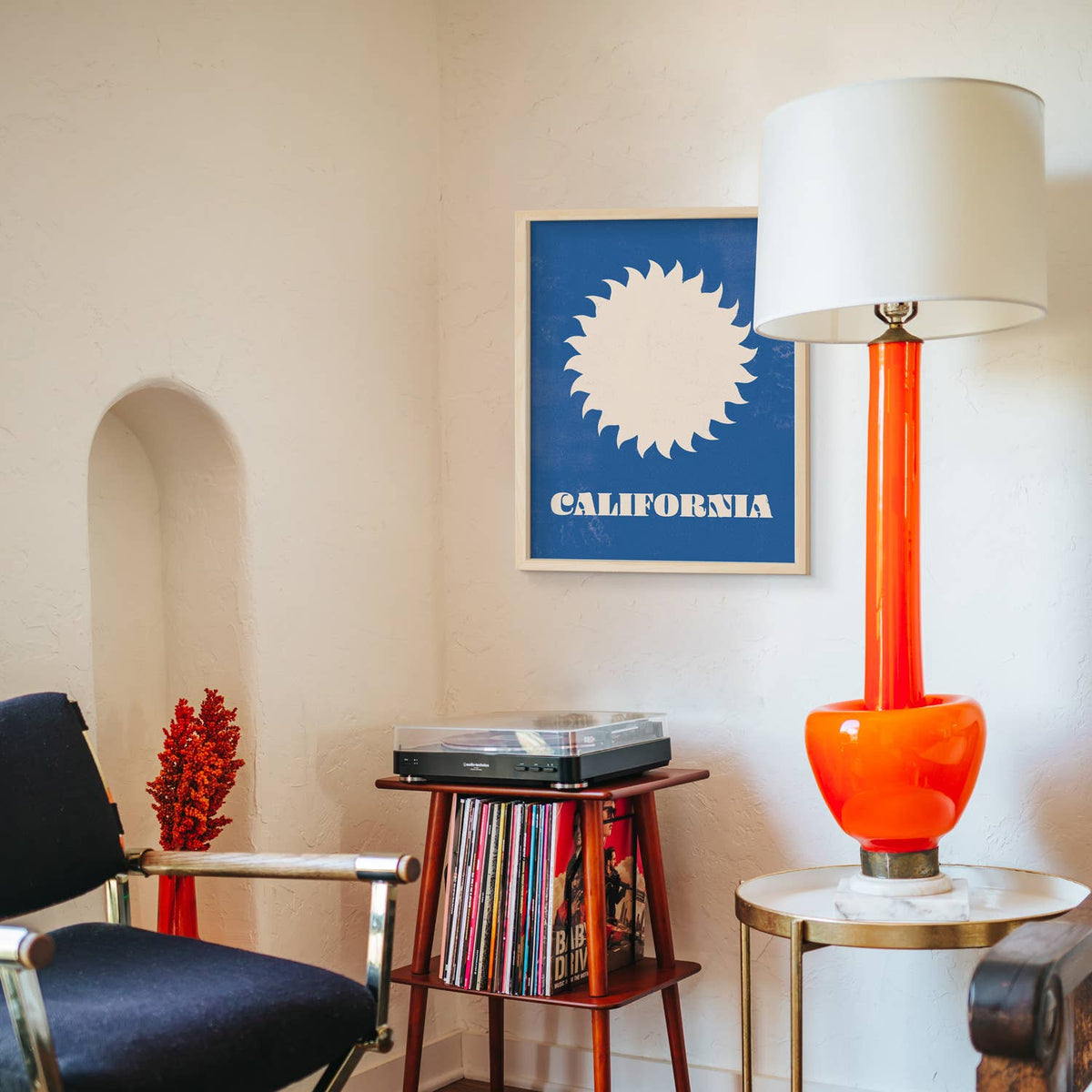A framed California poster with a blue background and white sunburst design hangs on a cream wall in a styled room. A tall orange table lamp with a white drum shade stands on a small round table beside it, and a record player sits on a wooden stand holding vinyl records.