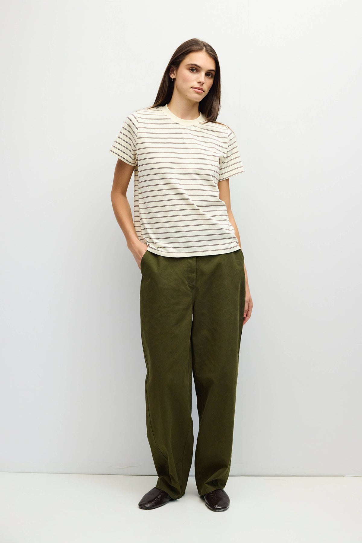 A woman modeling a short-sleeve crewneck t-shirt with thin beige horizontal stripes on a white background, paired with olive green wide-leg trousers and dark brown shoes.