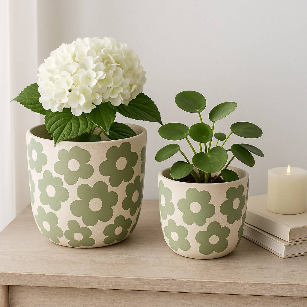 Two ceramic planters with a cream background and sage green flower pattern, styled on a light wood shelf with green plants, a white flower, and a lit candle.