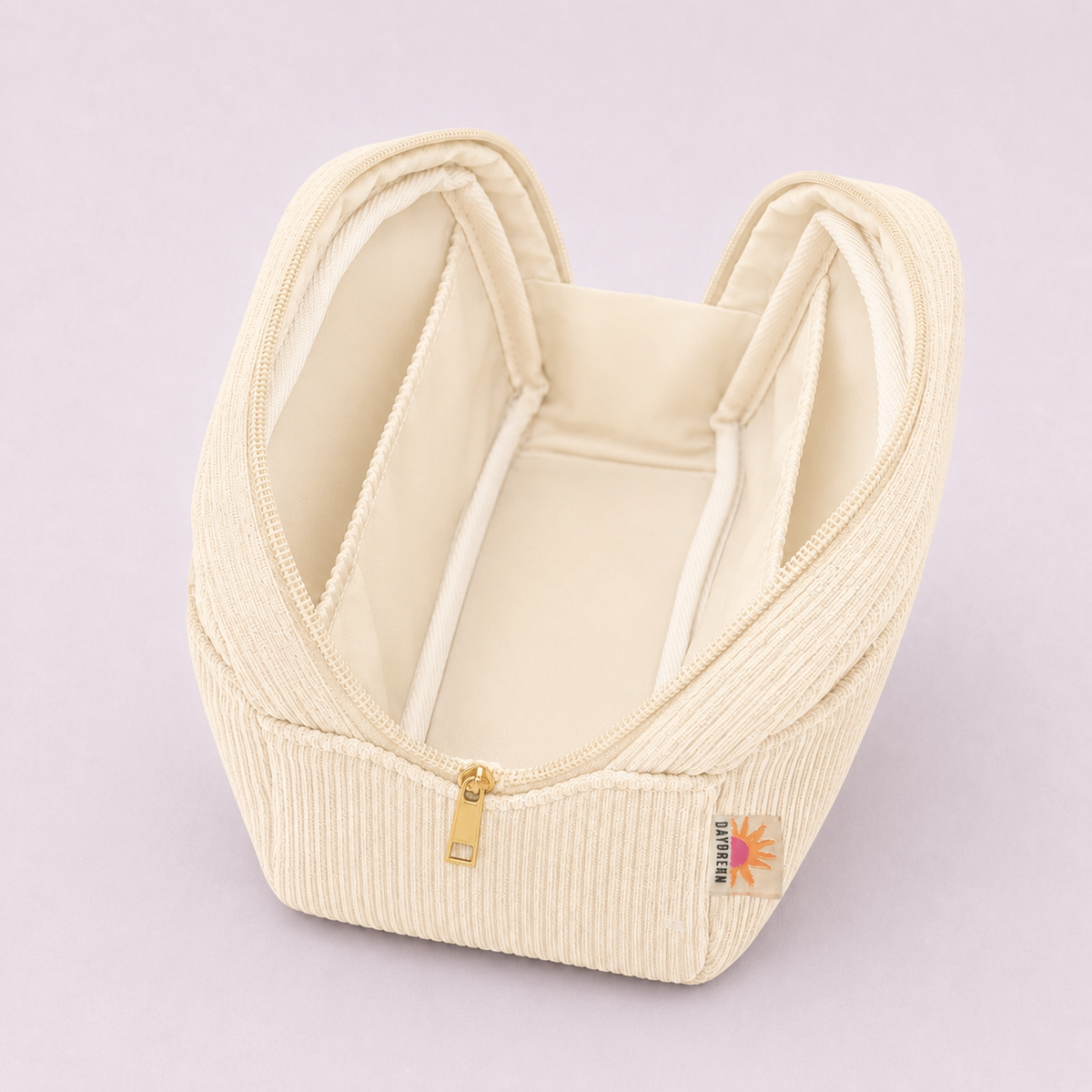 An off-white ribbed corduroy cosmetic bag with a gold zipper and a small colorful brand tag, shown on a light lavender background.
