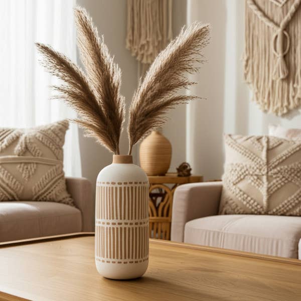 A cream ceramic vase with vertical ribbed detailing, filled with tall beige pampas grass, set on a wooden coffee table in a softly lit, modern boho living room.
