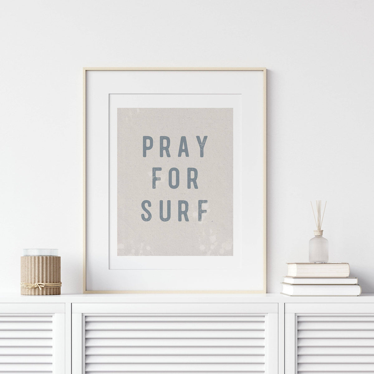 Framed artwork with 'PRAY FOR SURF' text on a white wall above a white cabinet.