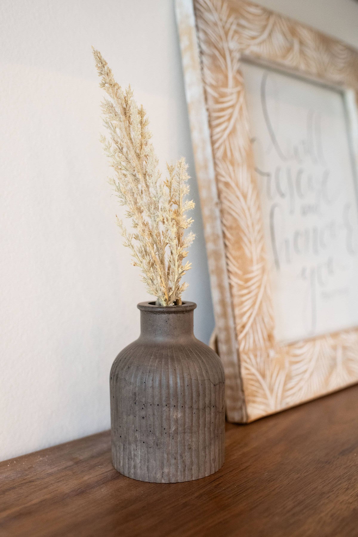 Decorative vase with dried plants on a wooden surface next to a framed picture.