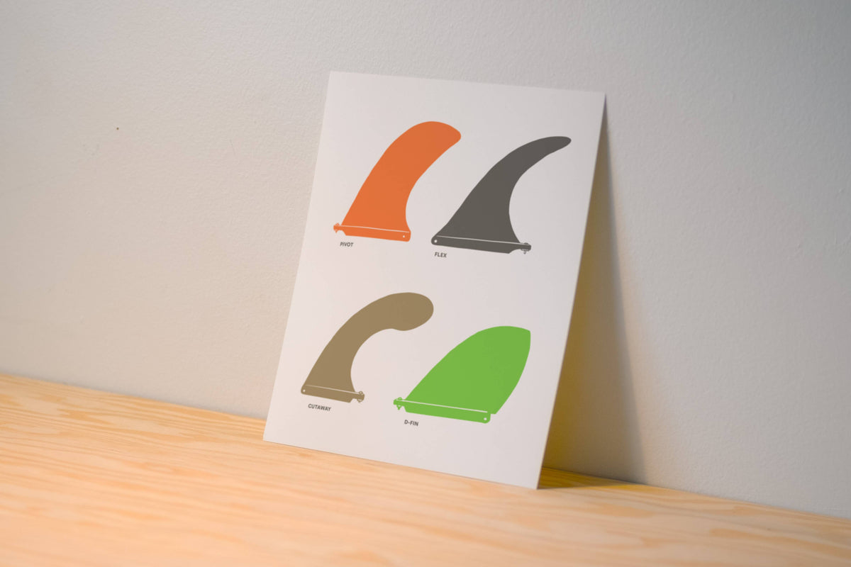 A vertical art print featuring four minimalist surfboard fin illustrations in orange, dark gray, tan, and green, displayed standing on a light wood surface against a neutral gray wall.